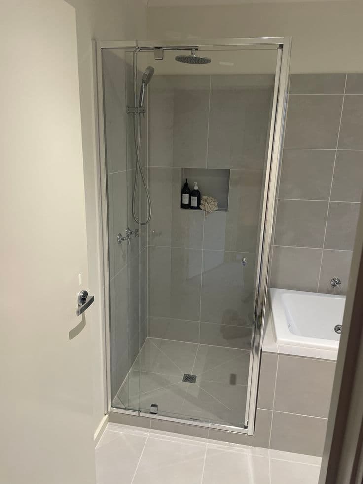 Semi-frameless shower enclosure with a chrome aluminium frame and pivot door in a grey-tiled bathroom with adjacent bathtub.