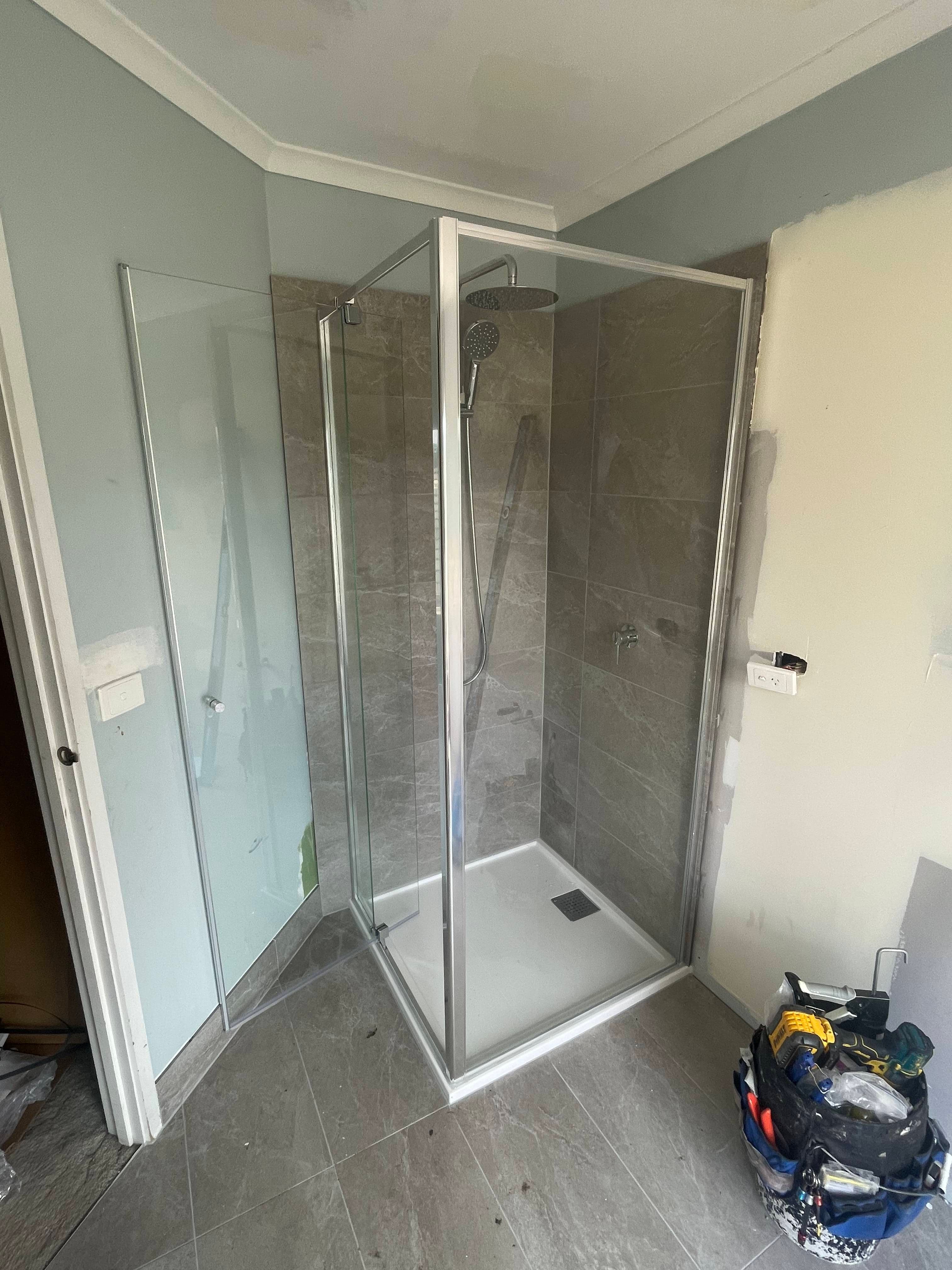 Semi-frameless shower enclosure with visible aluminium frame rails on all edges and a hinged door in a corner configuration, with chrome finish, set in a bathroom with grey marble-style tiles during renovation.