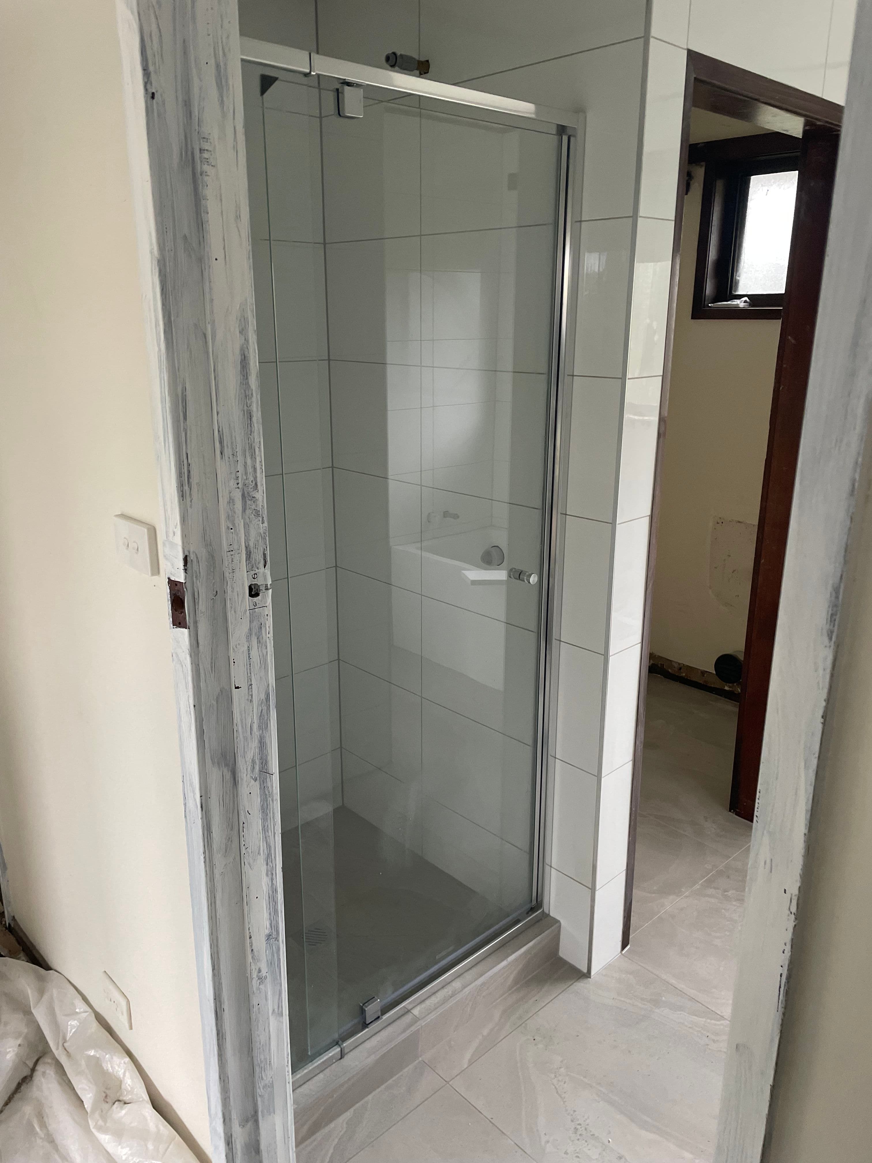 Semi-frameless shower screen with a single pivot door featuring visible chrome aluminium top rail and bottom channel in a white tiled bathroom under renovation.