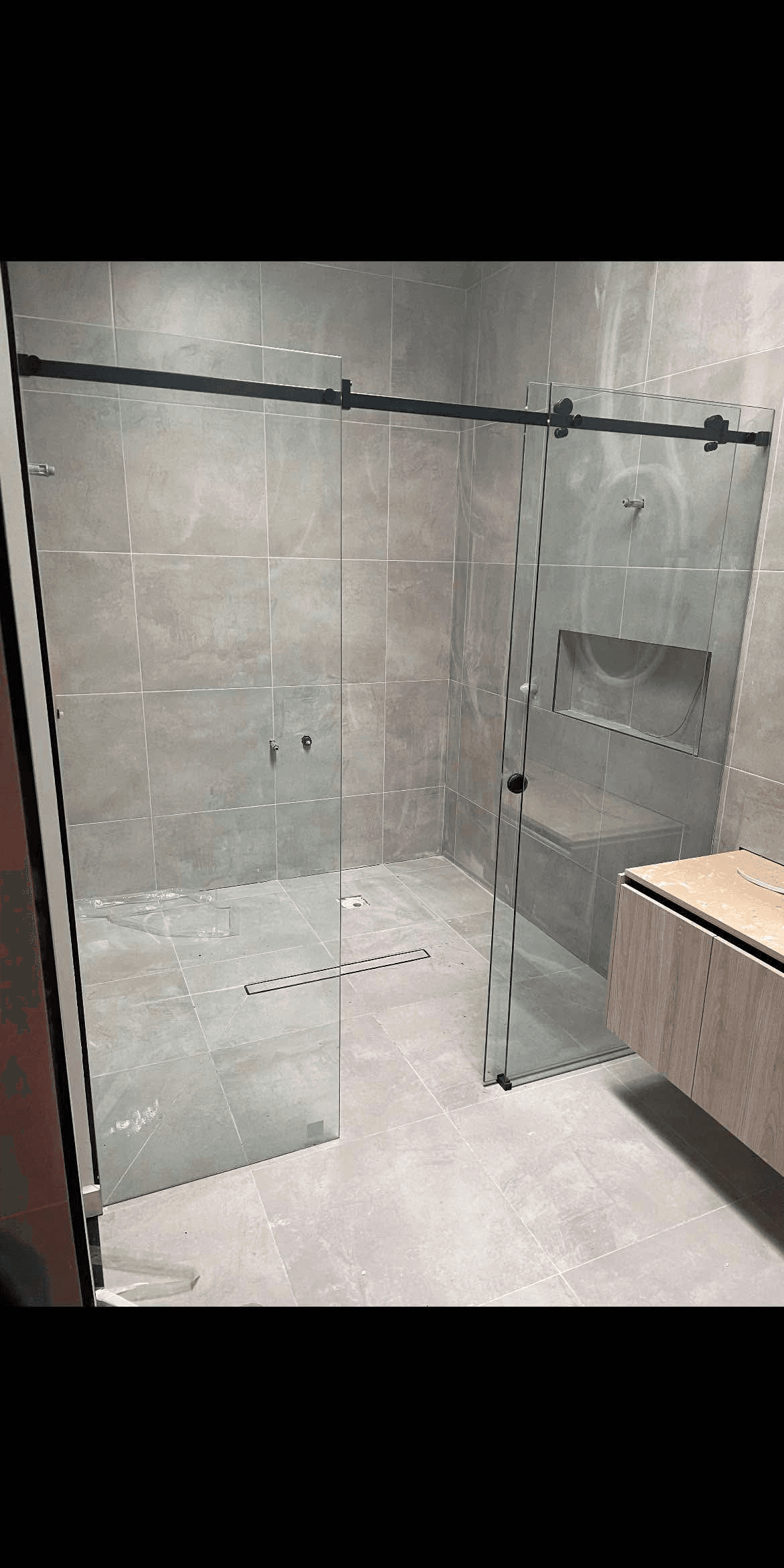 Semi-frameless shower screen with a matte black sliding door rail and fixed return panel in a large-format grey tiled bathroom with a built-in bench seat and timber vanity.