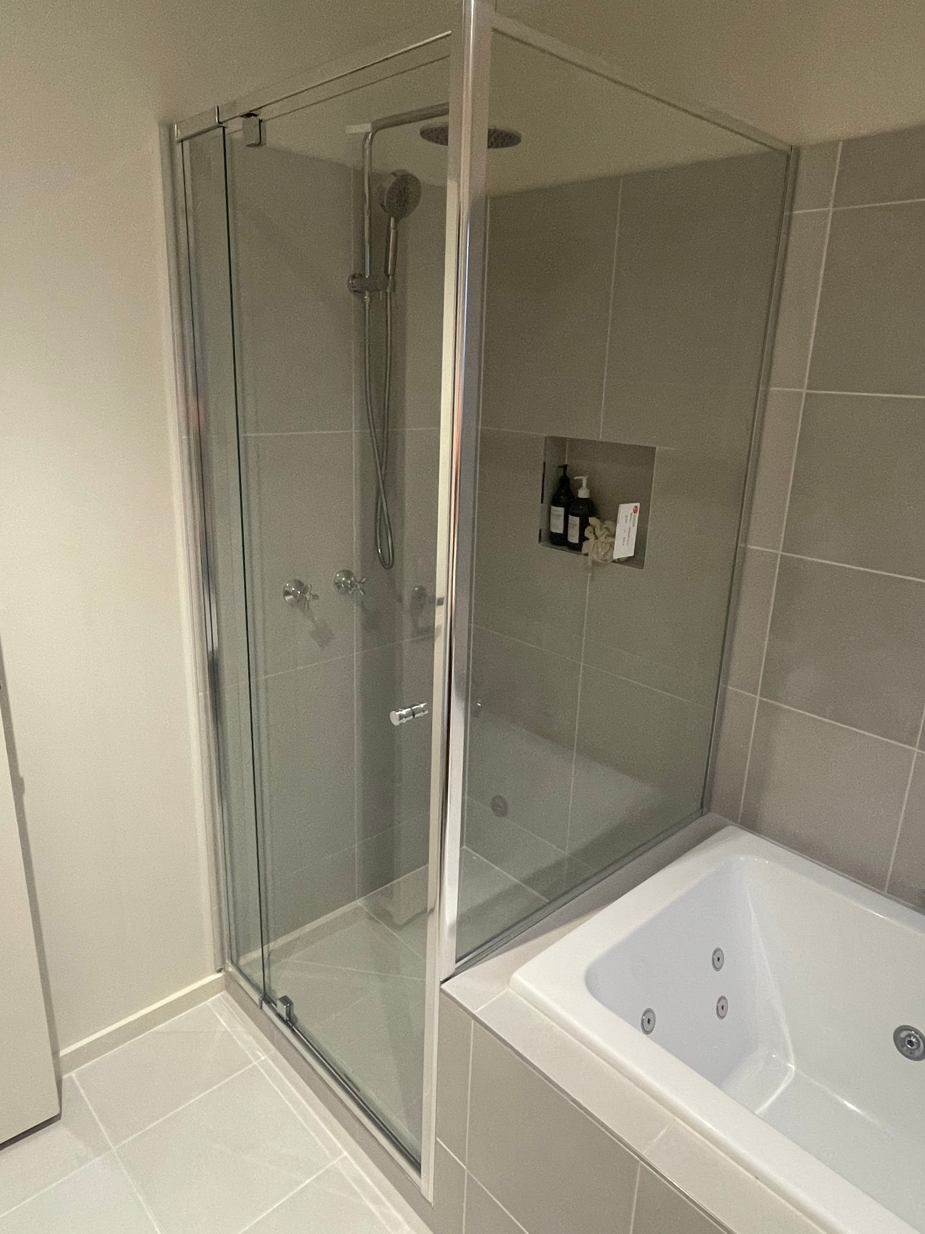 Semi-frameless shower enclosure with chrome aluminium frame rails, top channel, and base track forming a corner configuration adjacent to a freestanding spa bath in a grey-tiled bathroom.