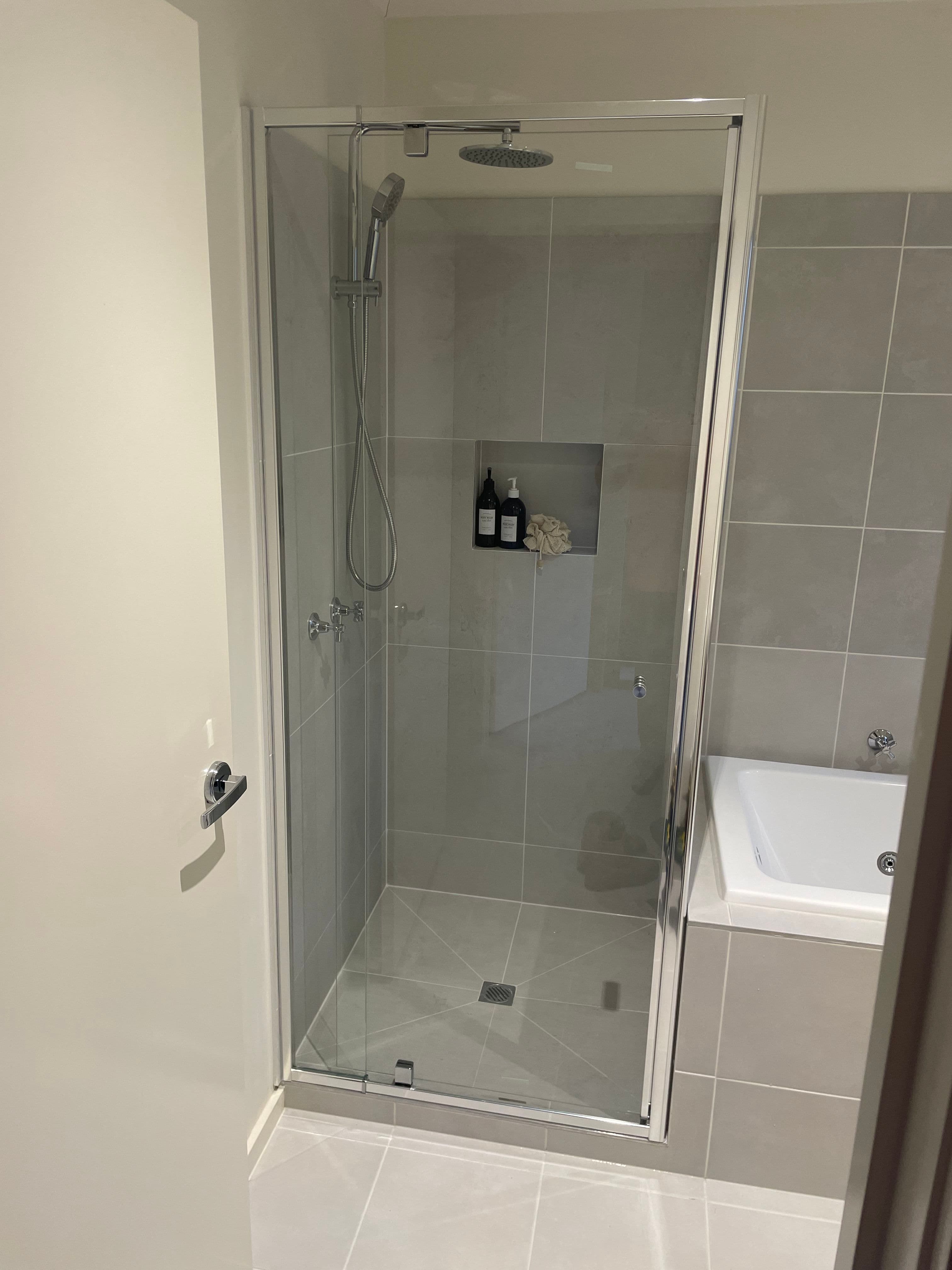 Semi-frameless shower enclosure with chrome aluminium frame rails on all sides and a hinged door in a grey-tiled bathroom with an adjacent freestanding bath.