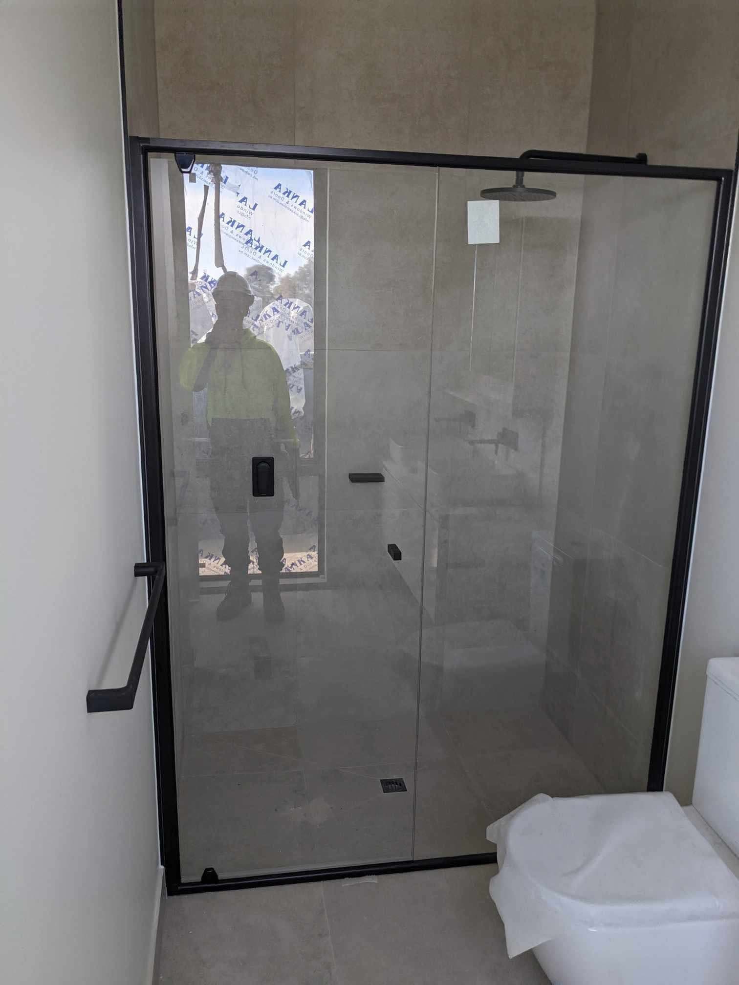 Semi-frameless shower screen with a sliding door configuration, matte black aluminium frame rails and track, clear glass, in a modern tiled bathroom with large format stone-look tiles.