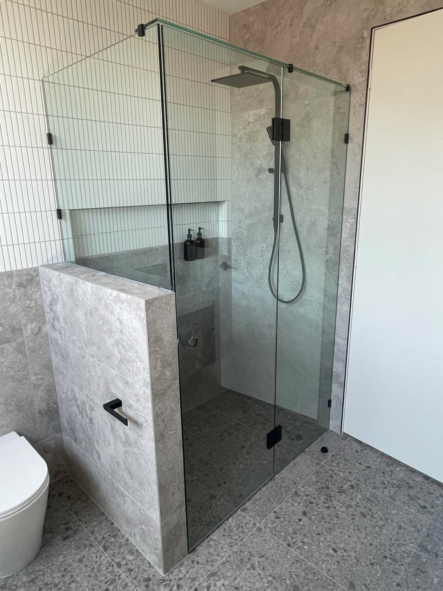 Frameless shower enclosure with matte black hinges and hardware in a corner configuration with a fixed return panel, featuring clear glass mounted directly on a tiled half-wall in a modern stone-tiled bathroom.