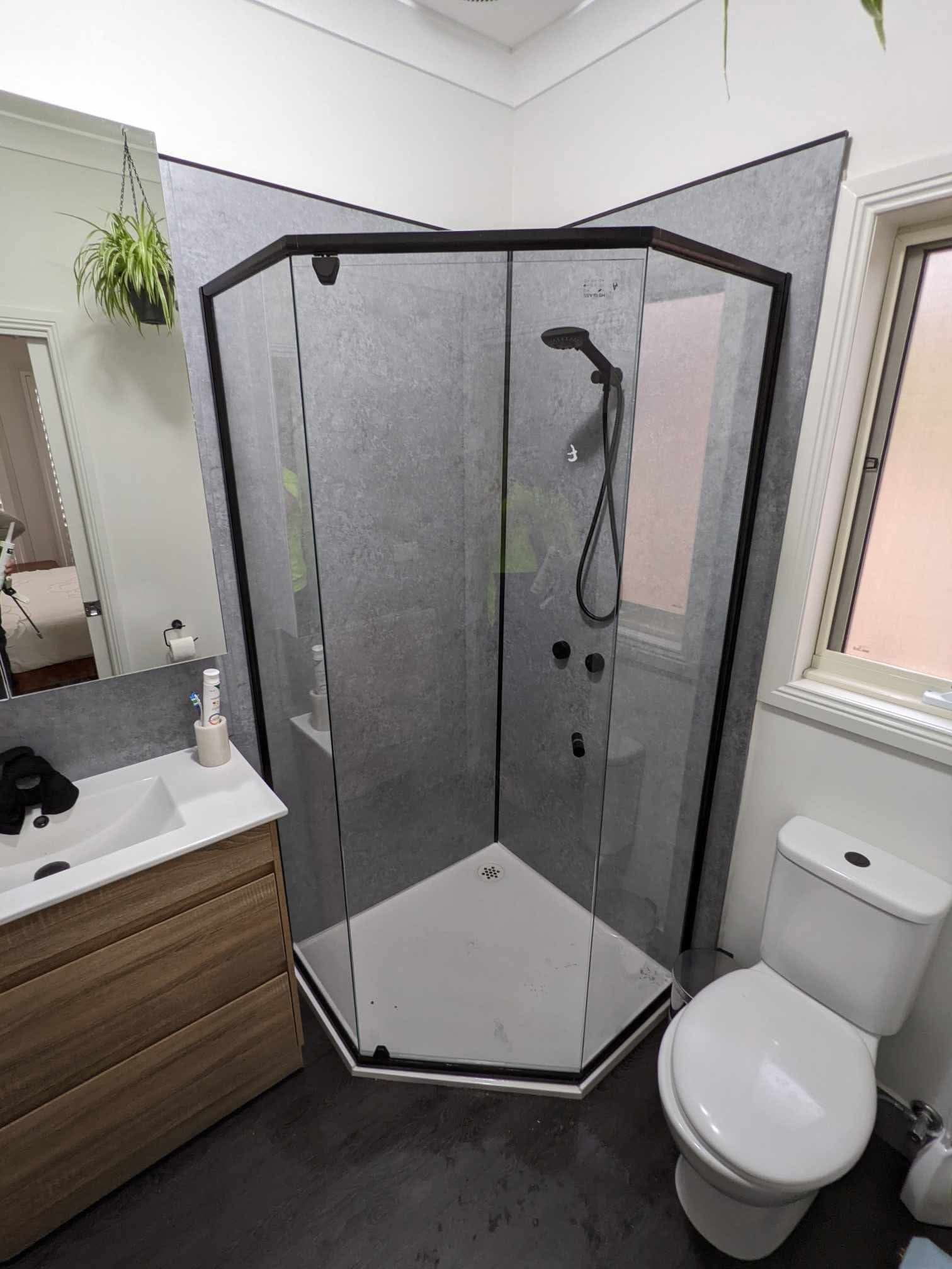 Semi-frameless neo-angle shower enclosure with matte black aluminium frame rails and a hinged door panel, set on a white shower base in a modern bathroom with grey stone-look wall panels.