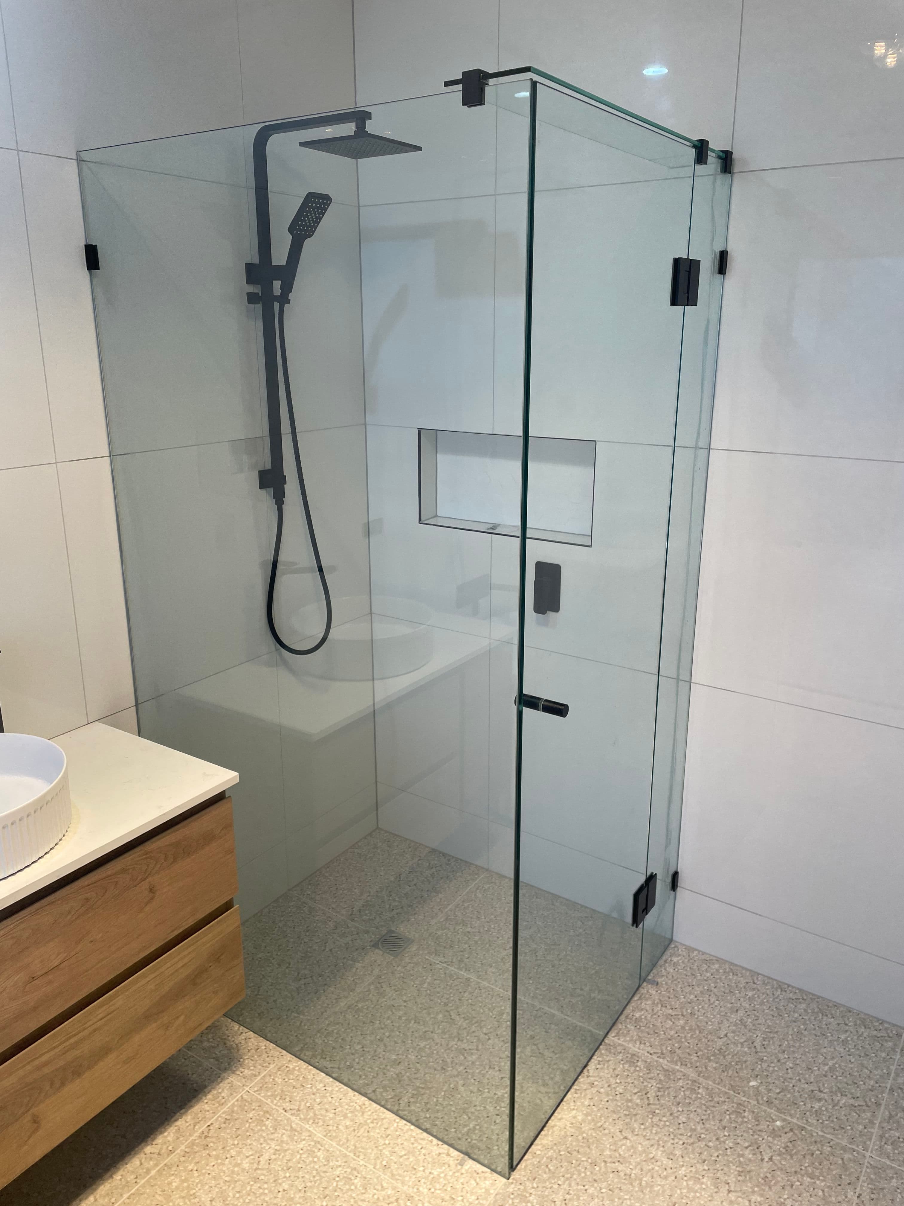 Frameless shower enclosure in a corner configuration with clear glass panels, matte black hinges and handle, featuring a hinged door and fixed return panel in a white tiled bathroom with terrazzo-style floor tiles.