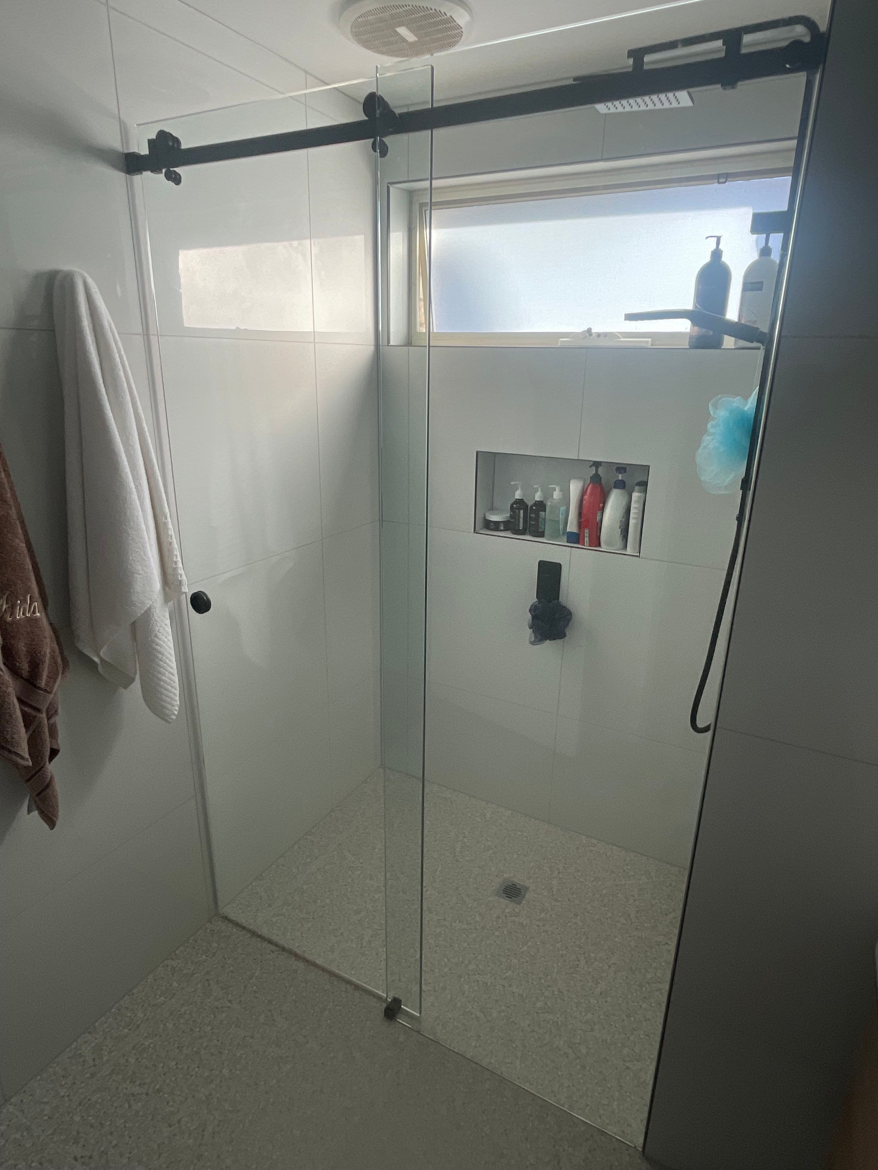 Semi-frameless shower enclosure with a sliding door on a matte black overhead barn-style track rail, featuring clear glass and a fixed side panel in a white tiled bathroom.