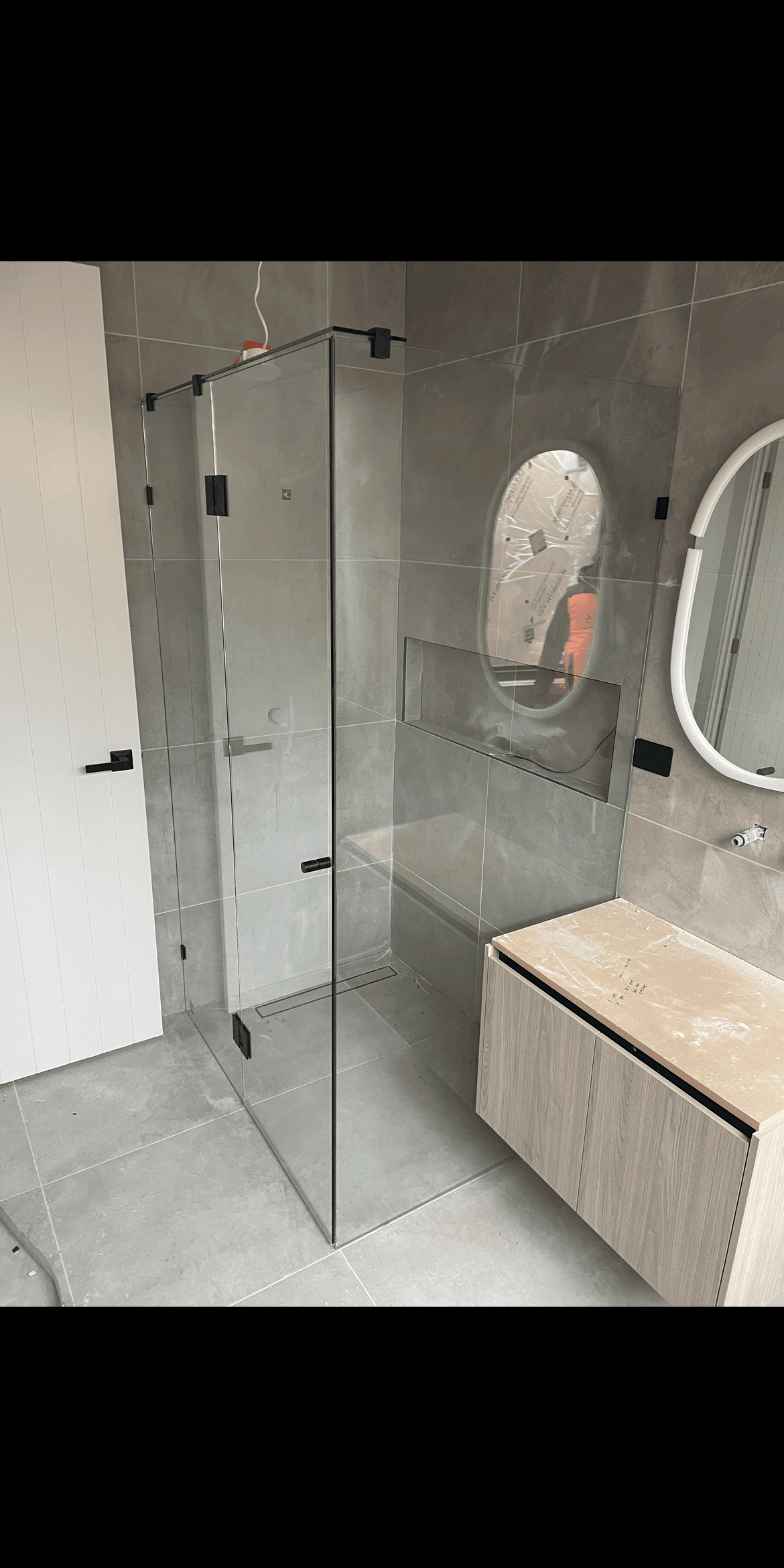 Frameless shower enclosure in a corner configuration with a hinged door, featuring matte black hinges and hardware screwed directly into the glass, set in a contemporary grey tiled bathroom with a floating timber vanity.