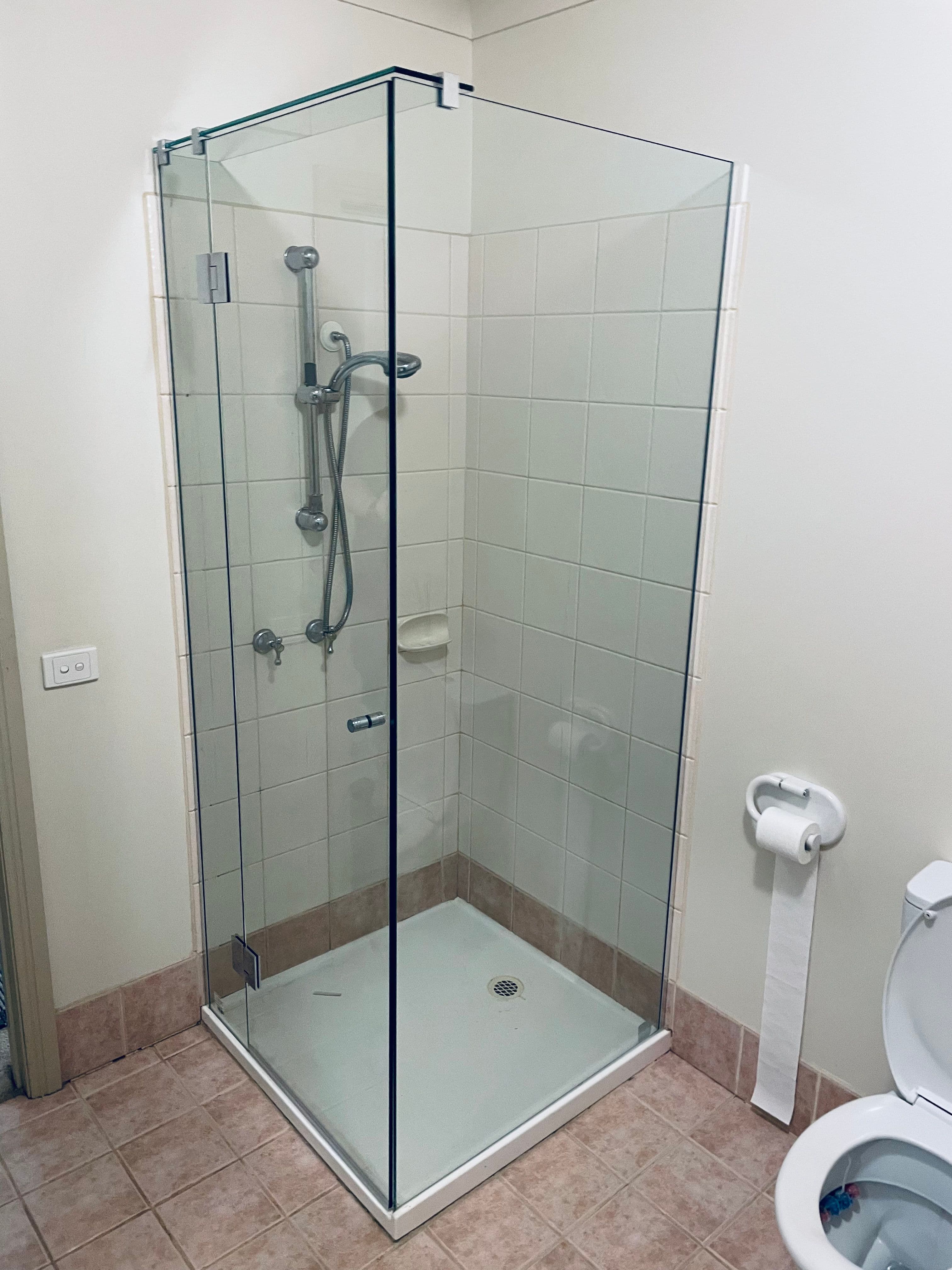 Semi-frameless shower enclosure with chrome hinges and a visible aluminium top rail and base channel in a corner configuration within a white-tiled bathroom.