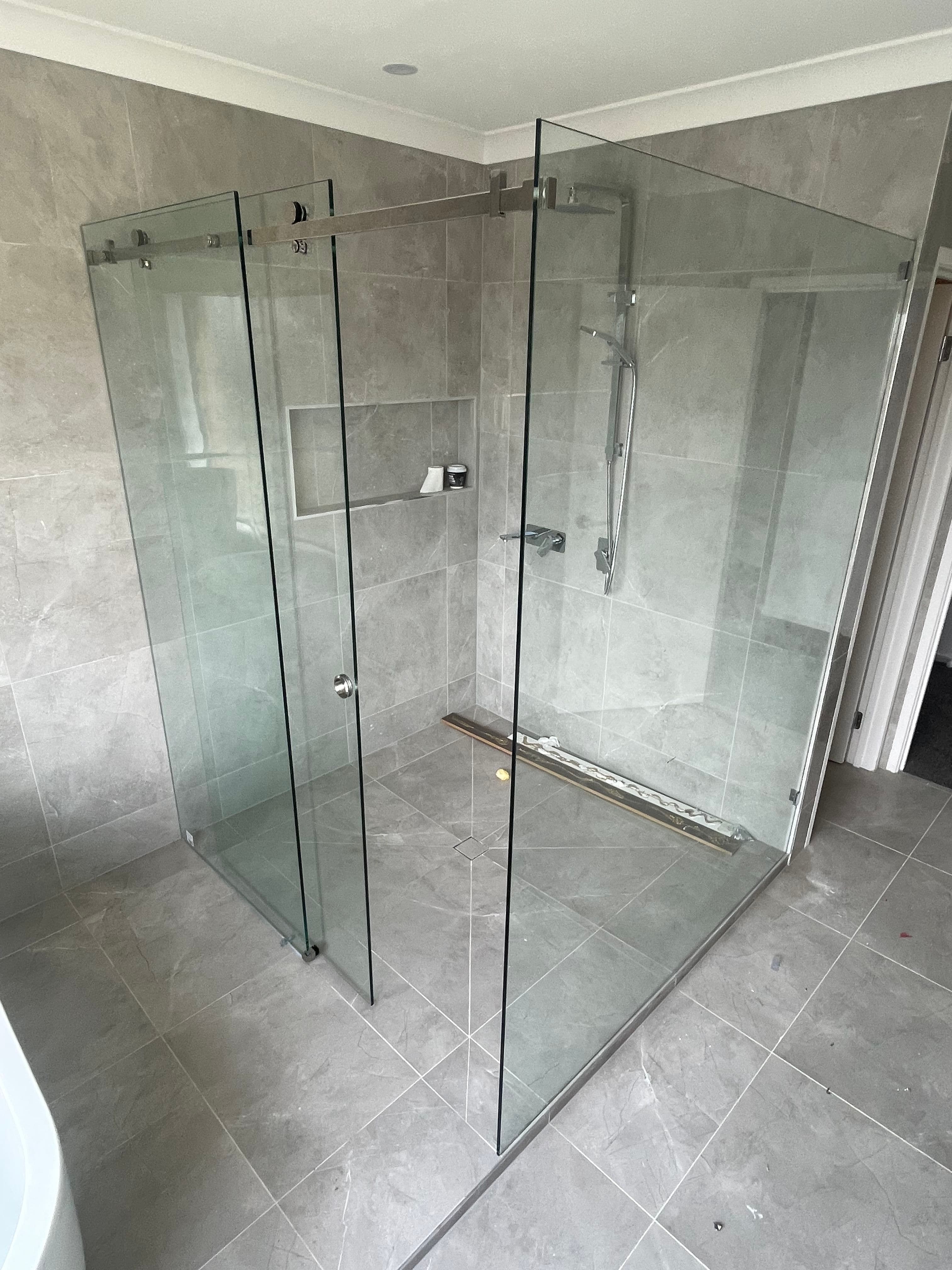 Semi-frameless corner shower enclosure with a sliding door on a visible top rail and brushed nickel hardware, featuring clear glass panels in a grey marble-tiled bathroom.