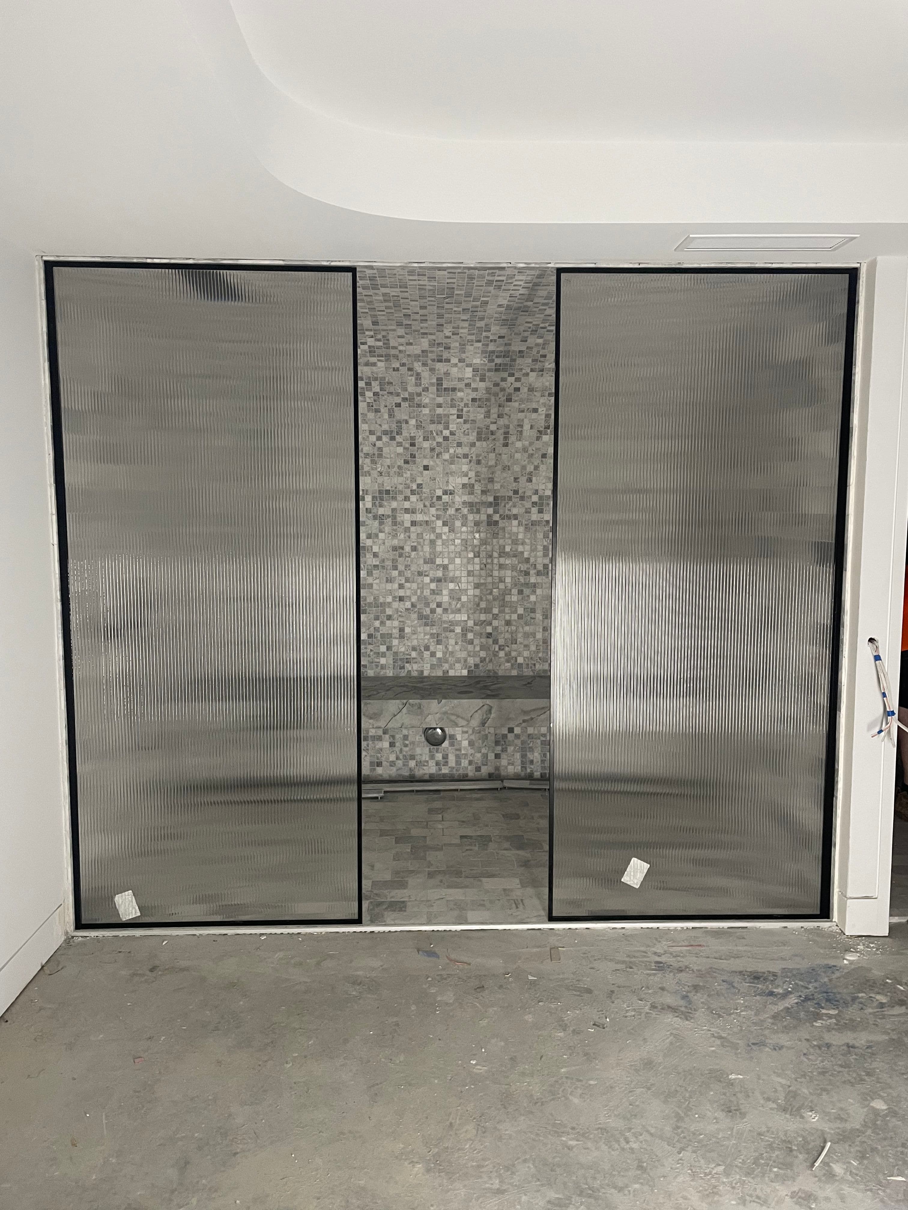 Semi-frameless shower enclosure with reeded glass panels set within a matte black aluminium frame channel, featuring a mosaic tile feature wall inside, photographed during construction.