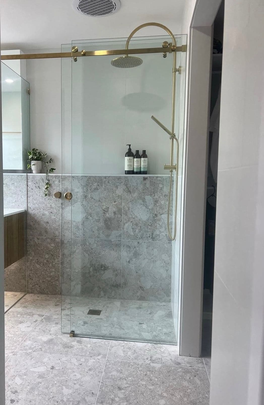 Semi-frameless shower screen with a brushed gold top rail and sliding door track in a marble-tiled ensuite with matching gold tapware and rainfall showerhead.