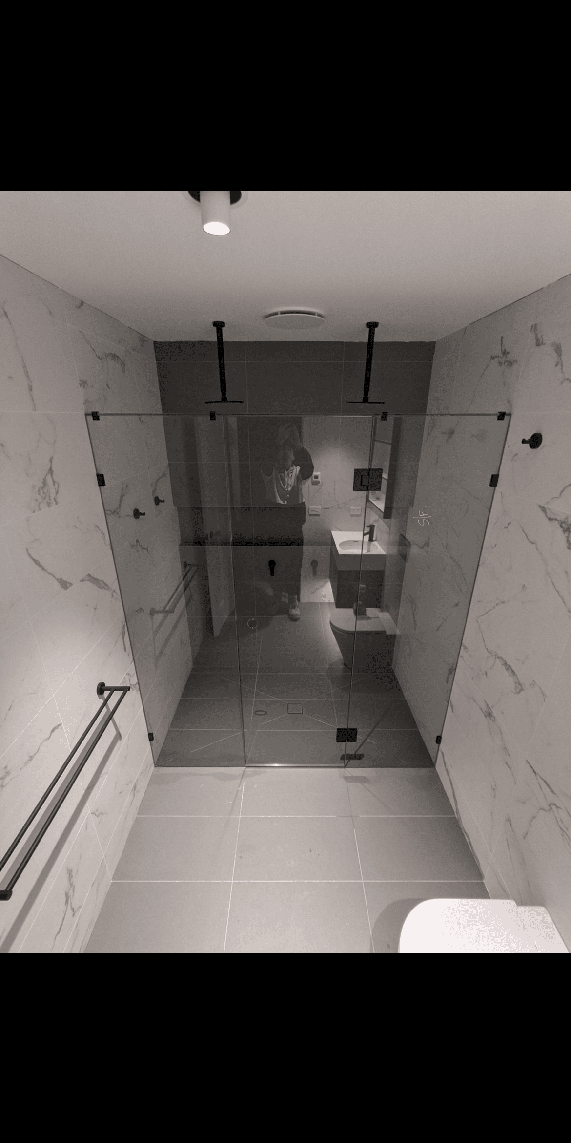 Frameless shower enclosure with matte black hinges and ceiling support bars in a corner configuration, set within a marble-tiled bathroom with large format grey floor tiles.