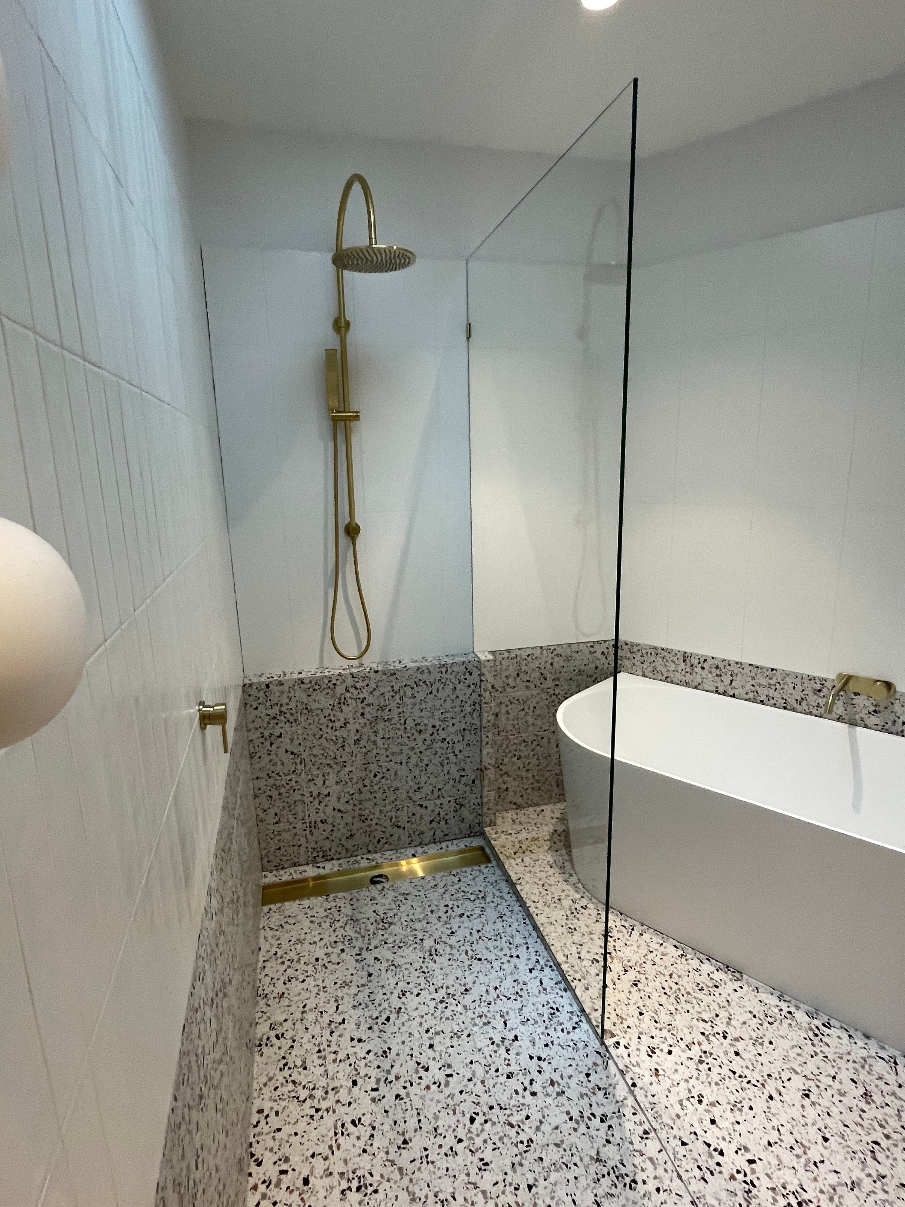 Frameless shower screen as a single fixed panel with brushed gold hardware, dividing an open wet room with terrazzo flooring, white tiled walls, a freestanding bath, and brushed gold shower fittings.