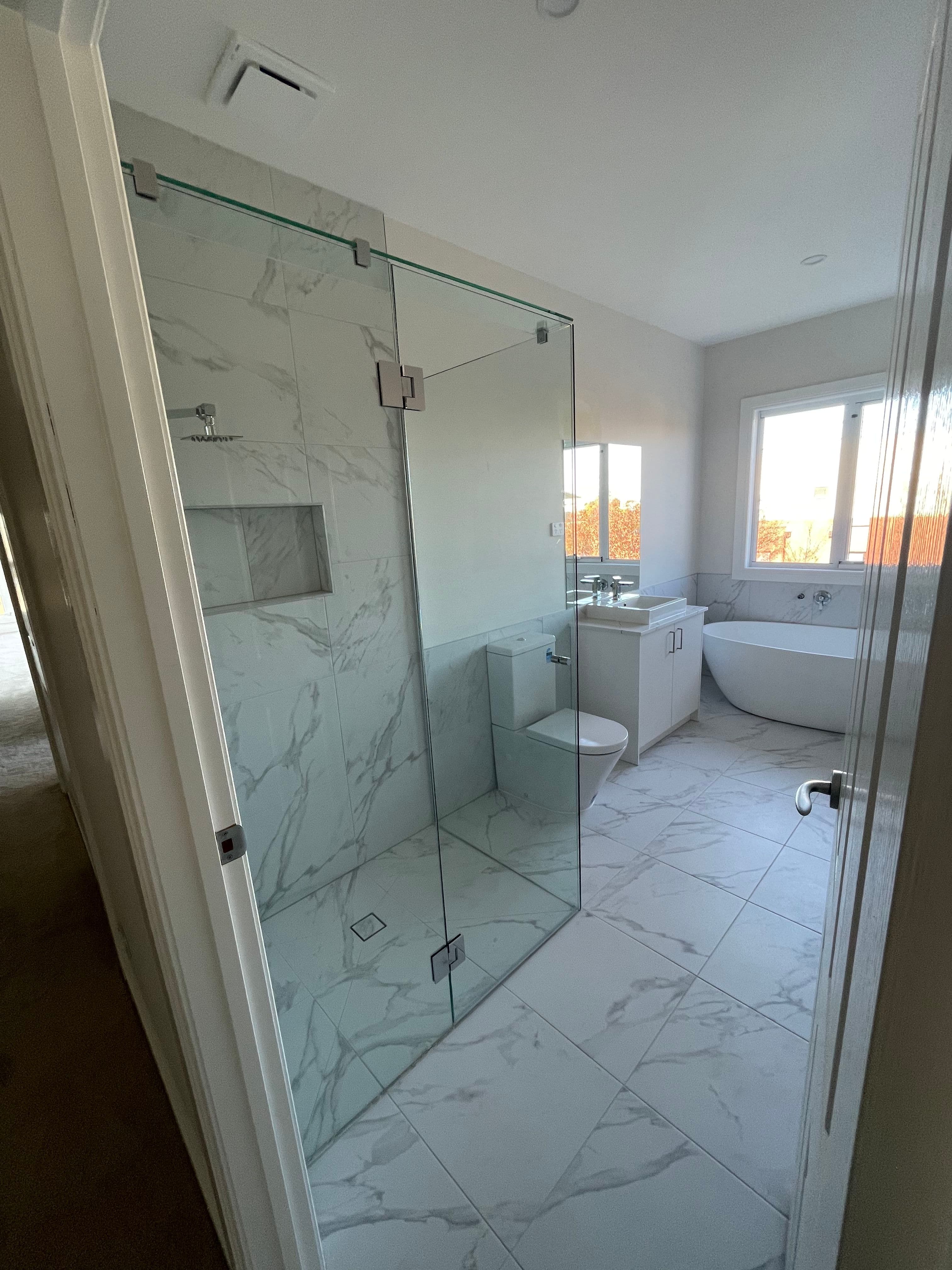 Frameless shower enclosure with clear glass panels in a return configuration, chrome hinges and clips mounted directly to the glass, set in a luxury marble-tiled ensuite with freestanding bath.
