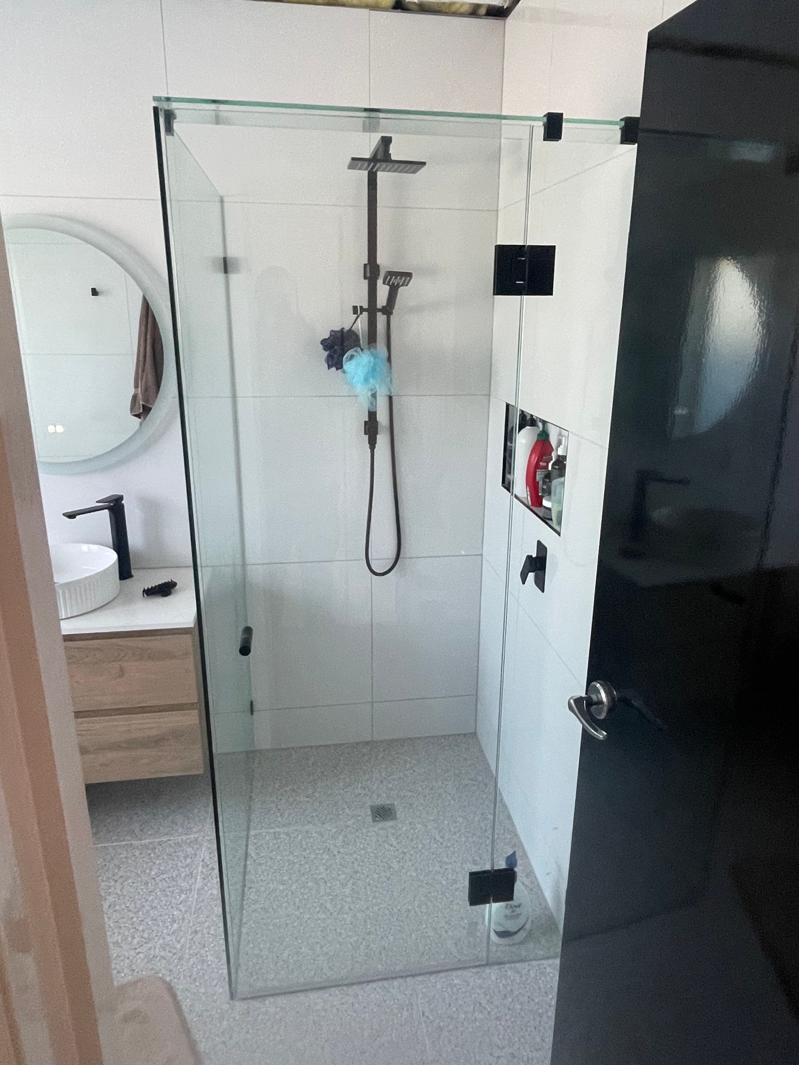 Frameless shower enclosure in a corner configuration with matte black hinges and clips mounted directly onto clear glass panels, set in a modern white-tiled bathroom with a timber vanity visible in the background.