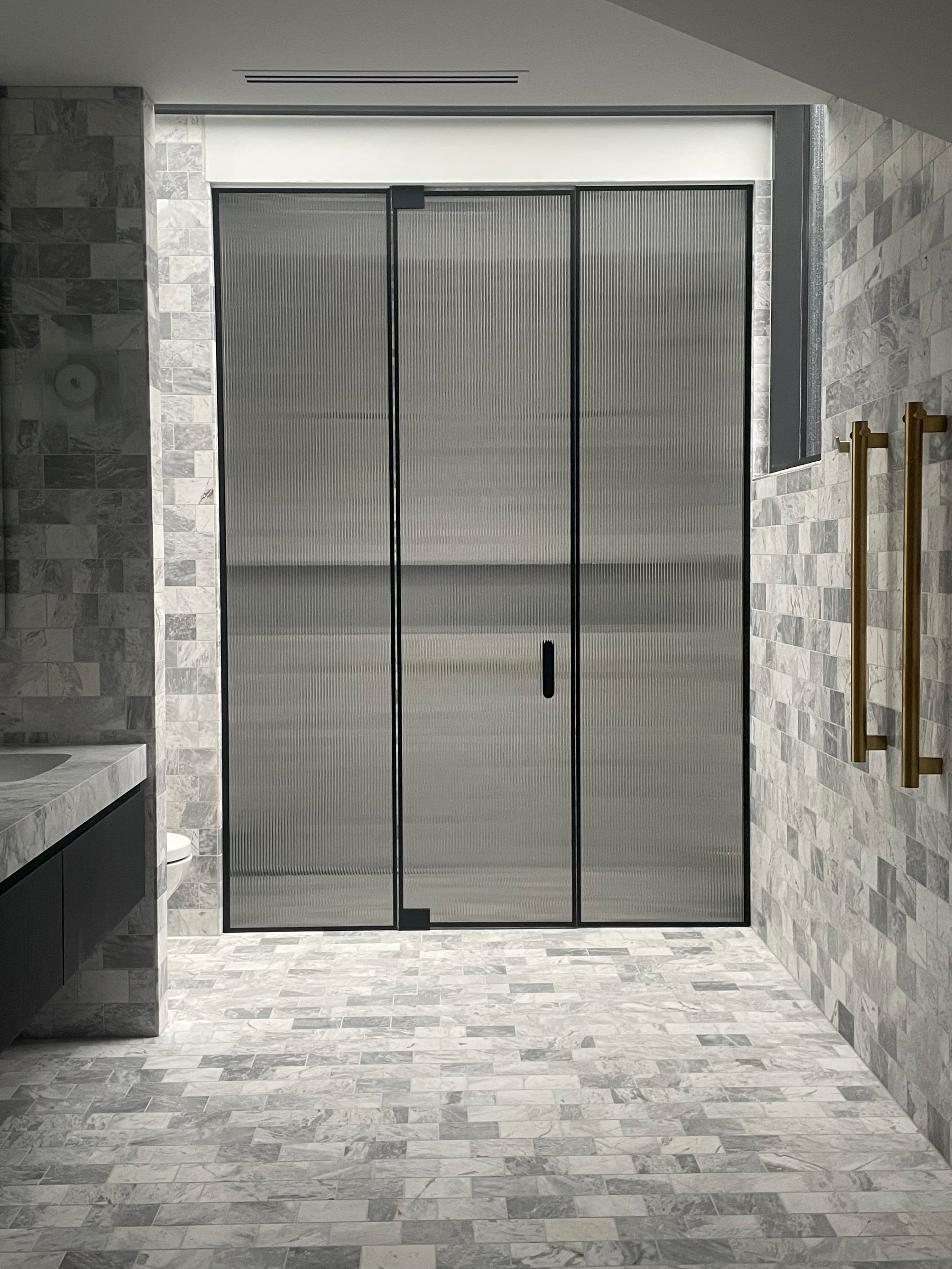 Semi-frameless shower enclosure with reeded glass panels in a three-panel sliding configuration, featuring matte black aluminium frame rails and a brushed gold towel bar in a marble-tiled bathroom.