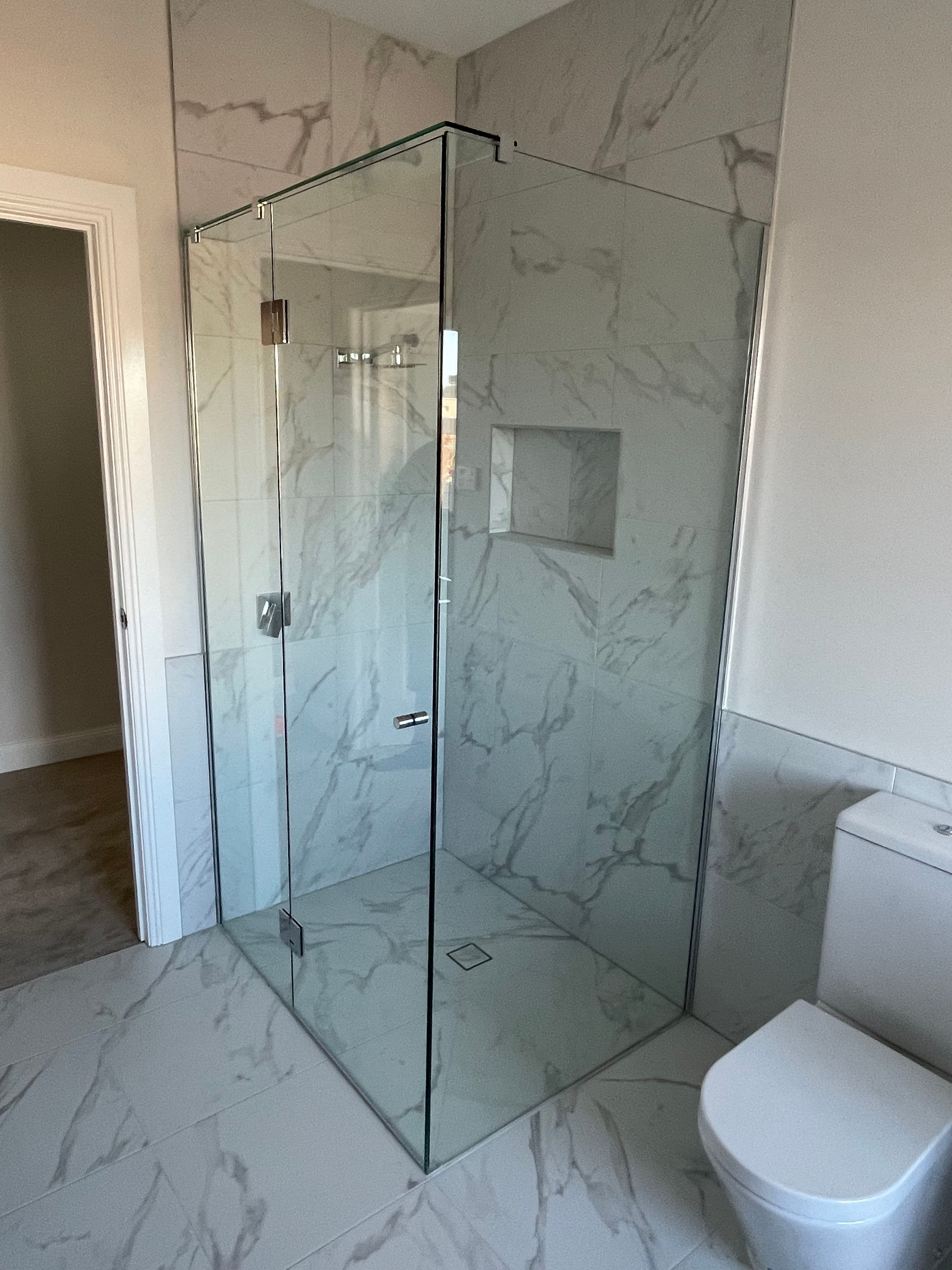 Frameless shower enclosure in a corner configuration with clear glass panels, chrome hinges and hardware, set in a luxurious marble-tiled bathroom with a square floor drain.