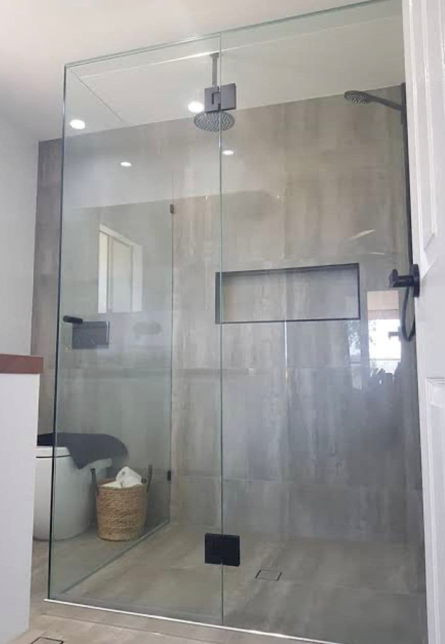 Frameless shower enclosure with matte black hinges and hardware in a corner configuration, featuring clear glass panels, a rain showerhead, and large-format grey tiles in a modern bathroom.