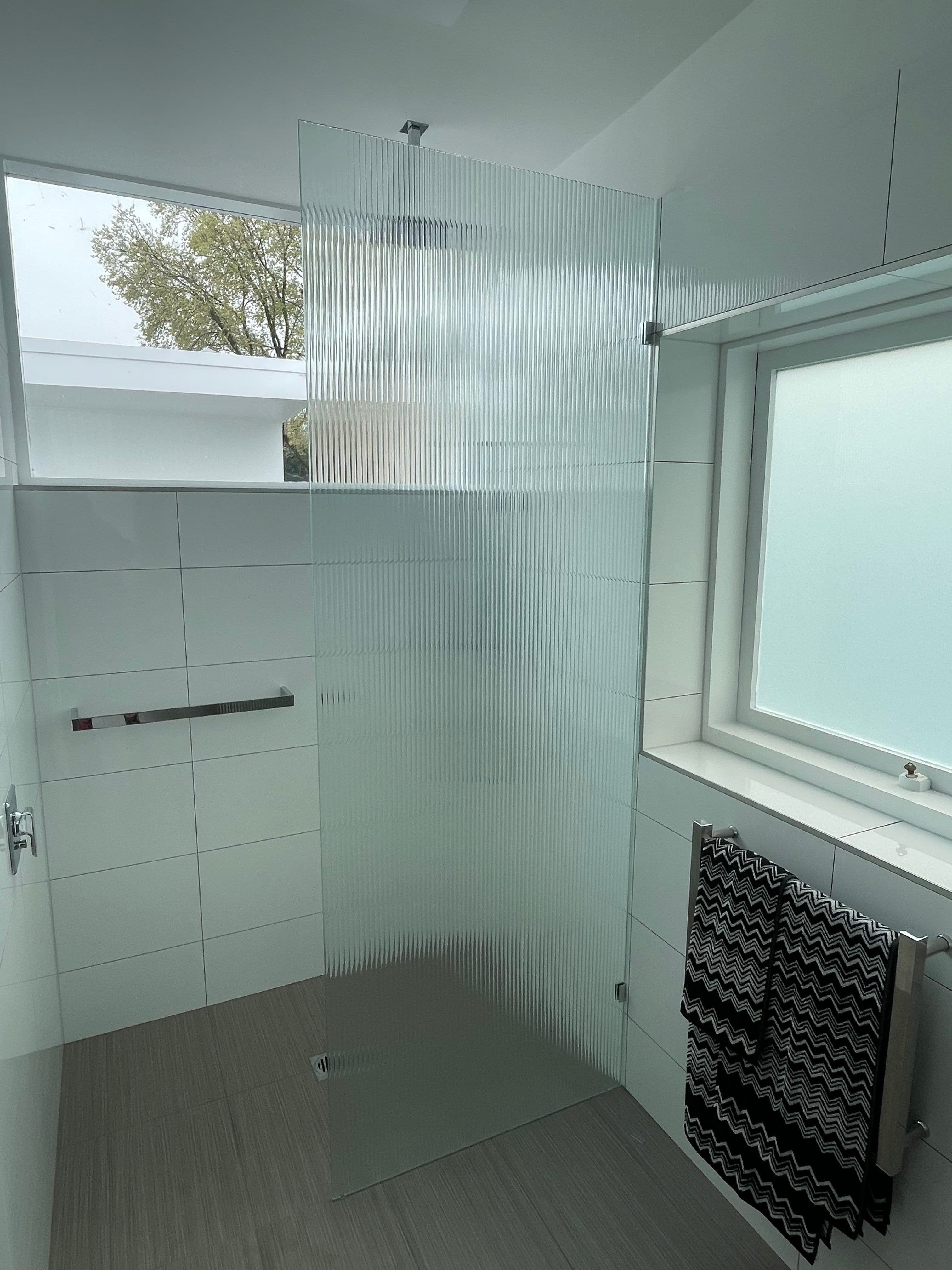 Frameless shower screen featuring reeded fluted glass panels in a corner configuration, fixed with chrome ceiling and floor clamps, in a modern white-tiled bathroom.