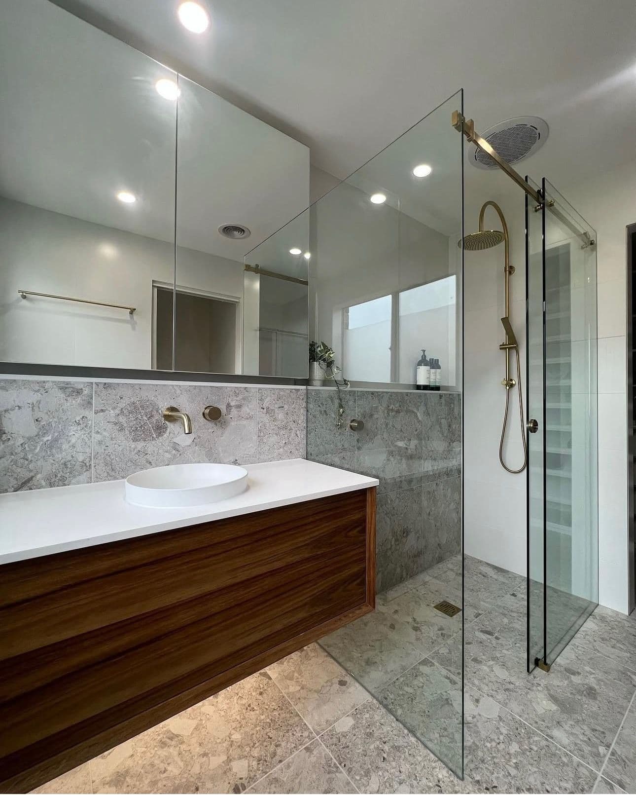 Semi-frameless shower enclosure with a brushed gold sliding door track and hardware in a modern bathroom featuring marble-look tiles, timber vanity, and vessel sink.