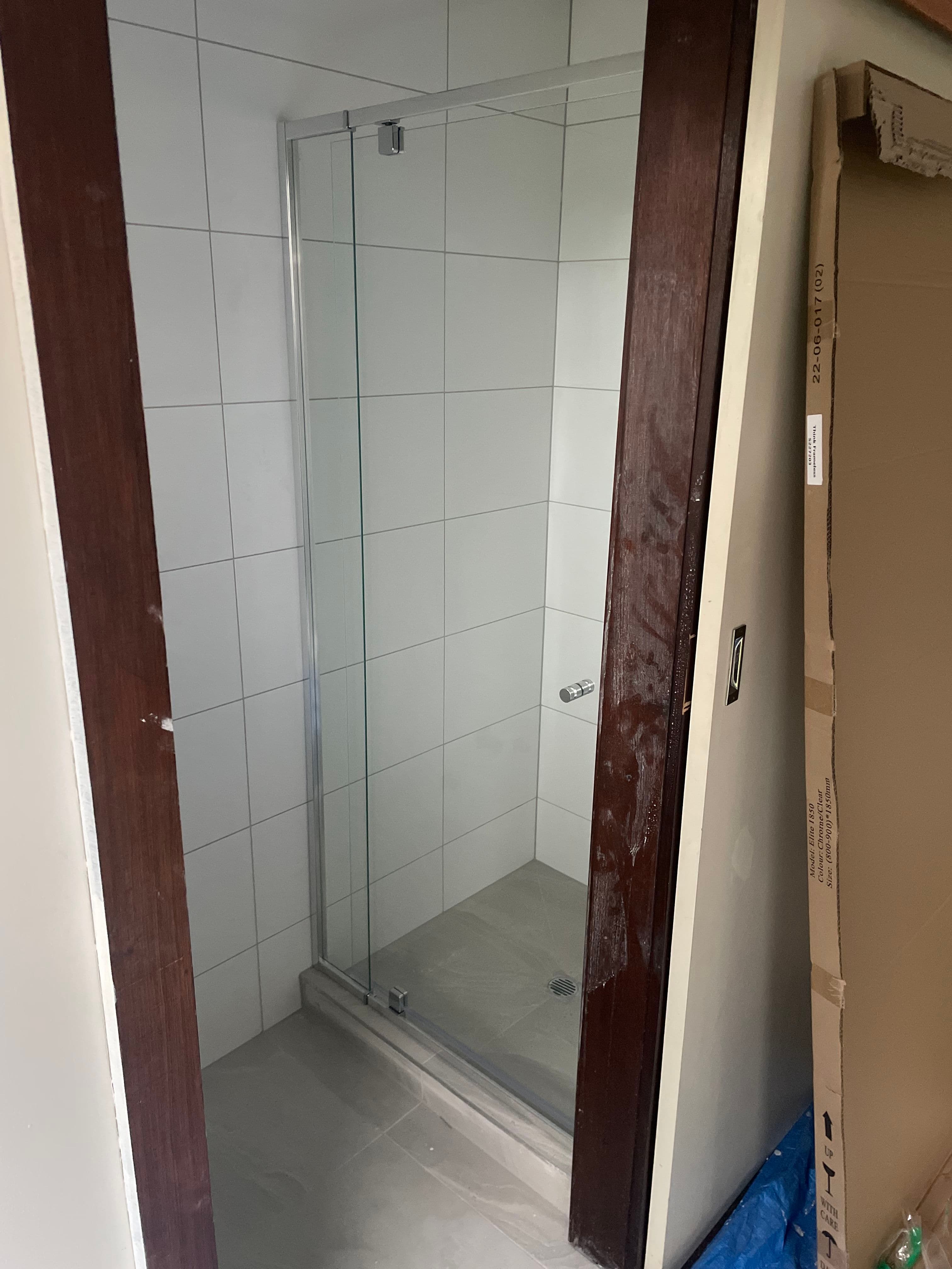 Semi-frameless shower screen with a chrome aluminium top rail and bottom track, single pivot door configuration in a white tiled bathroom under construction.