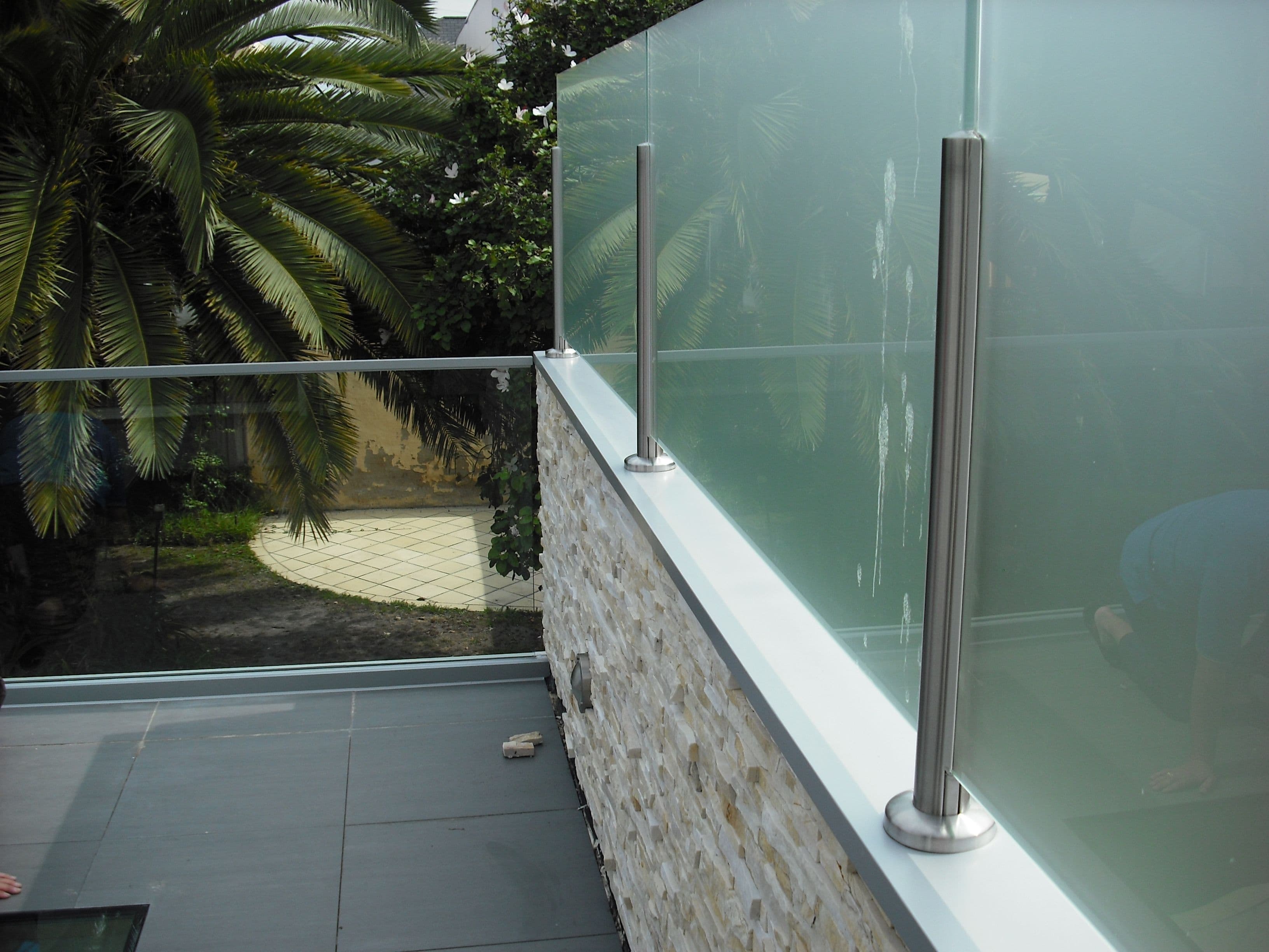Glass balustrade with brushed nickel post spigots mounted on a stone-clad wall edge of an elevated outdoor terrace with slate tile flooring, overlooking a garden.