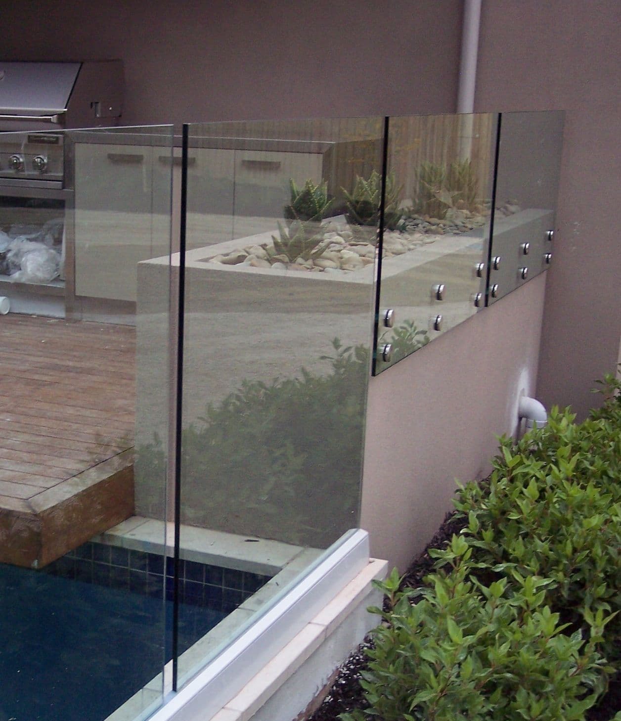 Frameless glass pool fence with chrome standoff fixings mounted on a rendered wall, surrounding a tiled swimming pool in a backyard alfresco setting.