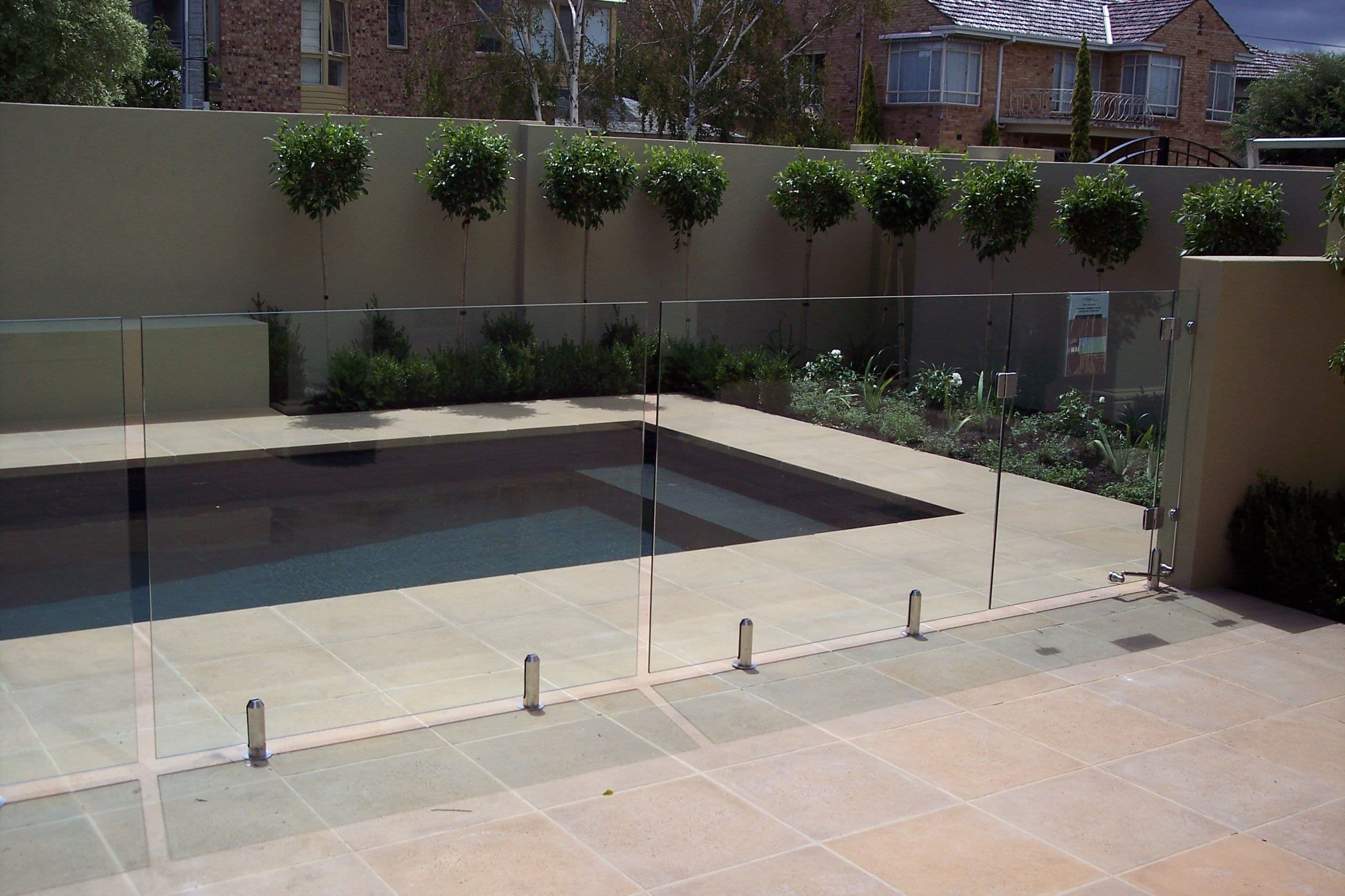 Frameless glass pool fence with chrome spigots core-drilled into sandstone coping surrounding a backyard swimming pool in a landscaped suburban courtyard.