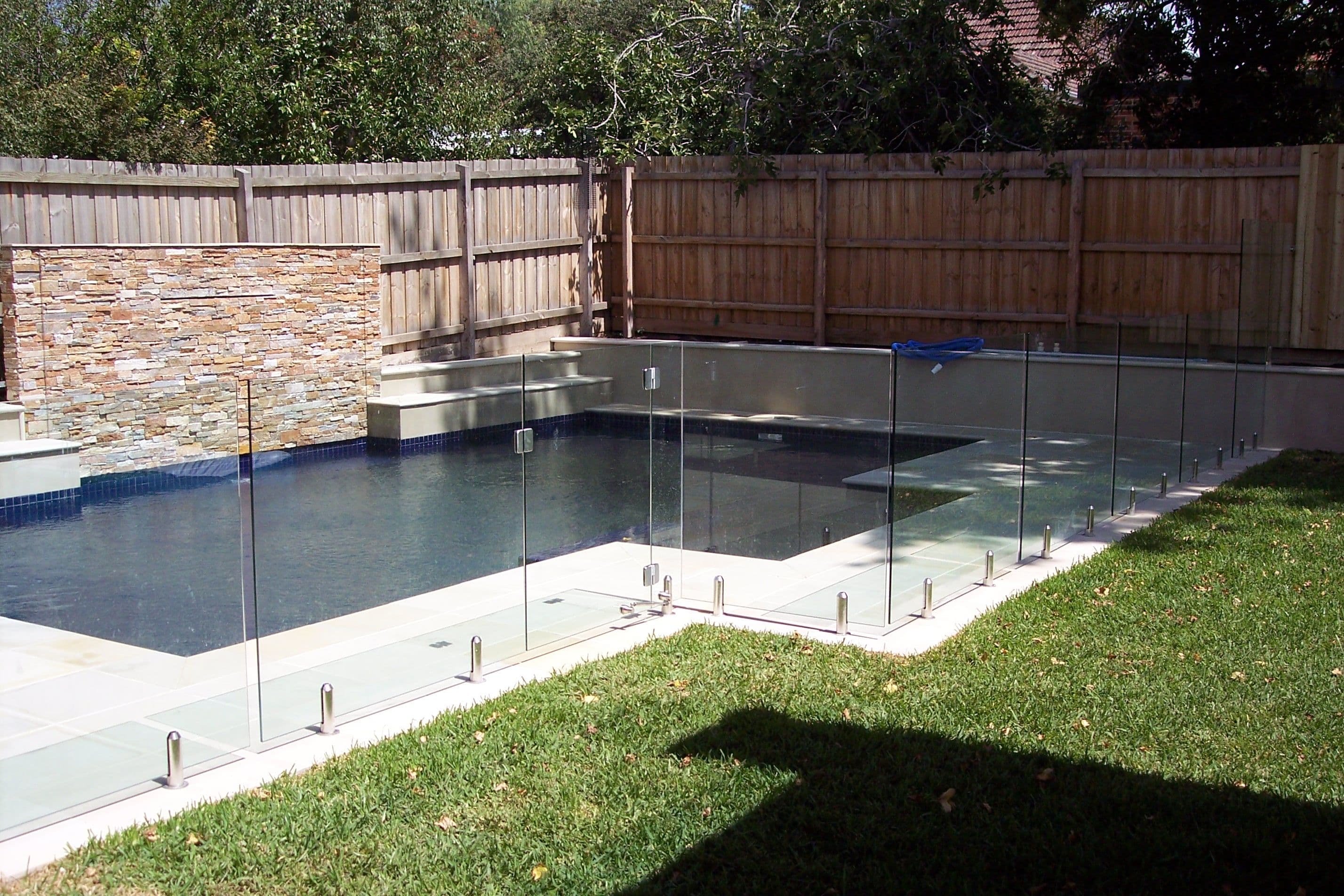 Frameless glass pool fence with chrome spigots core-drilled into concrete coping surrounding a residential backyard pool with a stone feature wall.