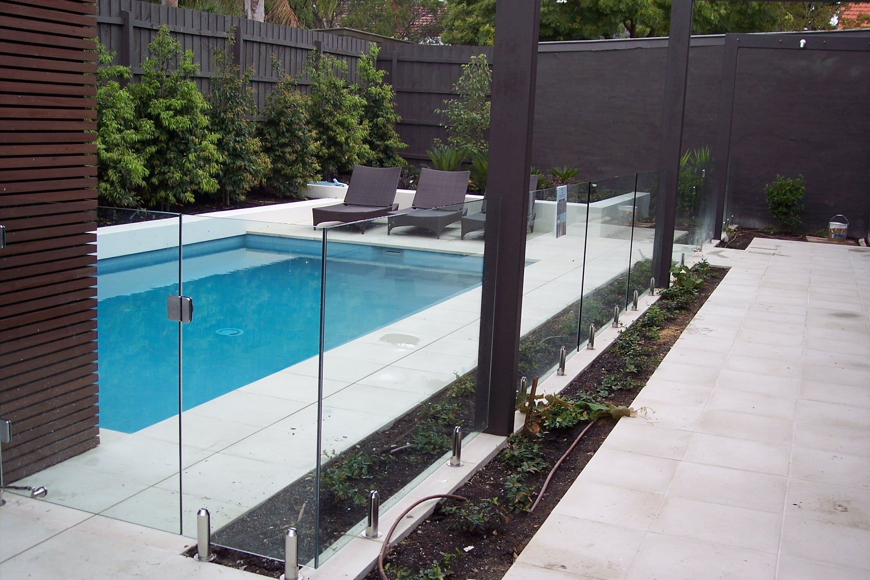 Frameless glass pool fence with chrome spigots core-drilled into concrete coping surrounding a rectangular backyard swimming pool in a modern outdoor setting.