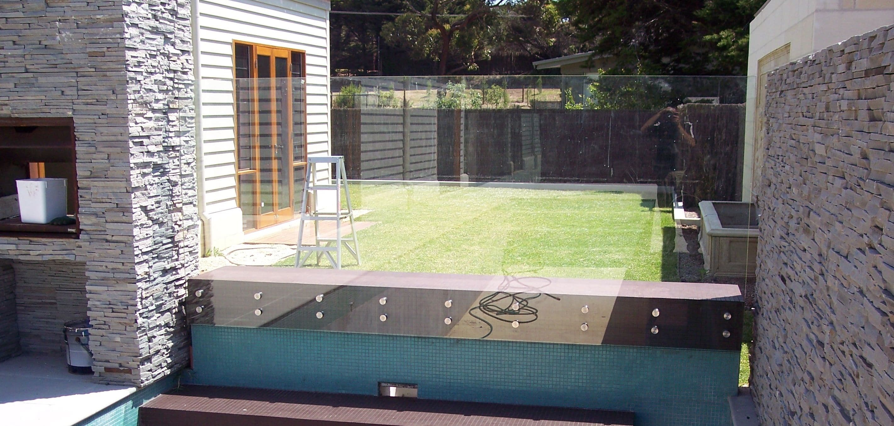 Frameless glass pool fence with chrome spigots mounted on the coping of an elevated pool surround, with teal mosaic tile pool visible in the foreground and backyard lawn beyond.