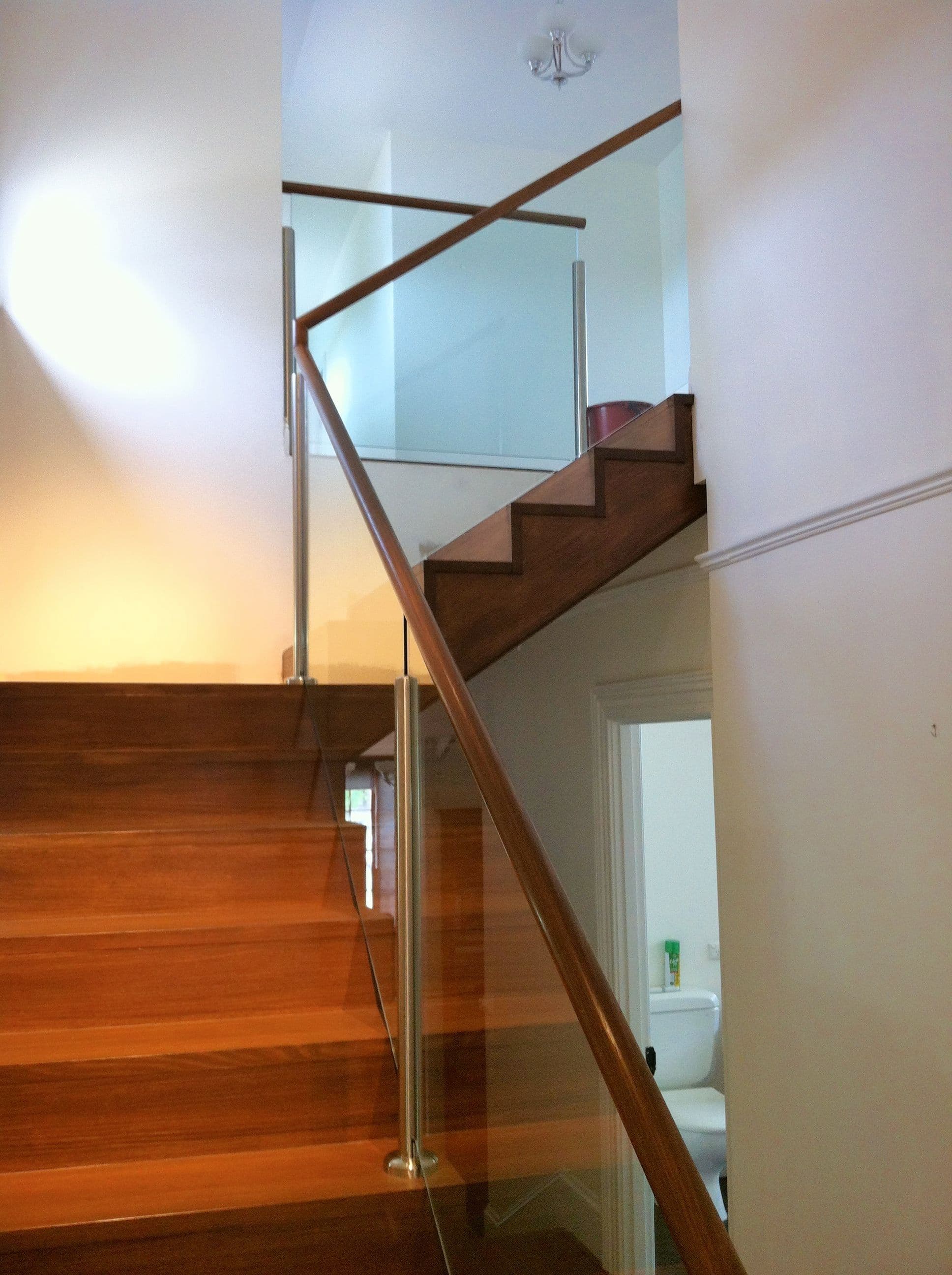 Clear glass stair balustrade with chrome spigots and timber handrail alongside timber staircase in a residential interior.