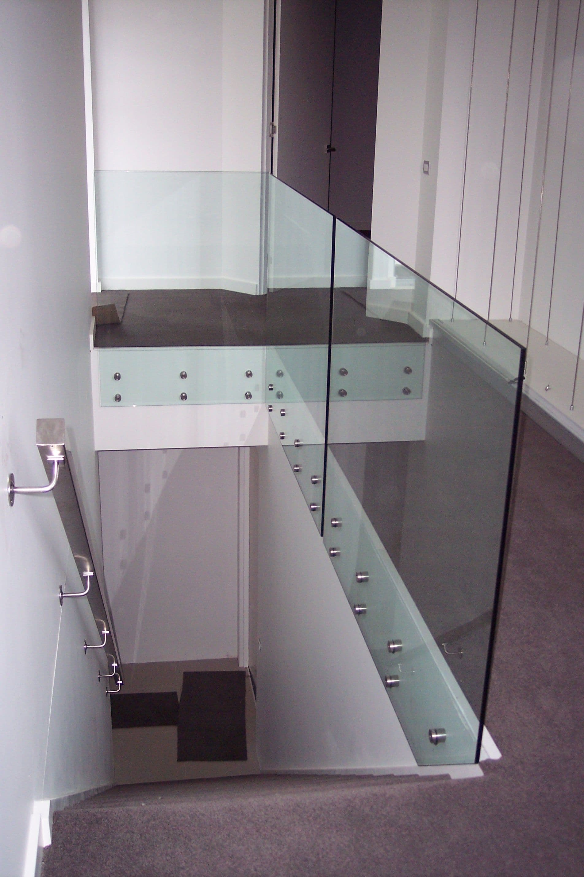 Clear glass staircase and landing balustrade with chrome standoff fixings mounted to a base channel, viewed from above in a modern interior stairwell.