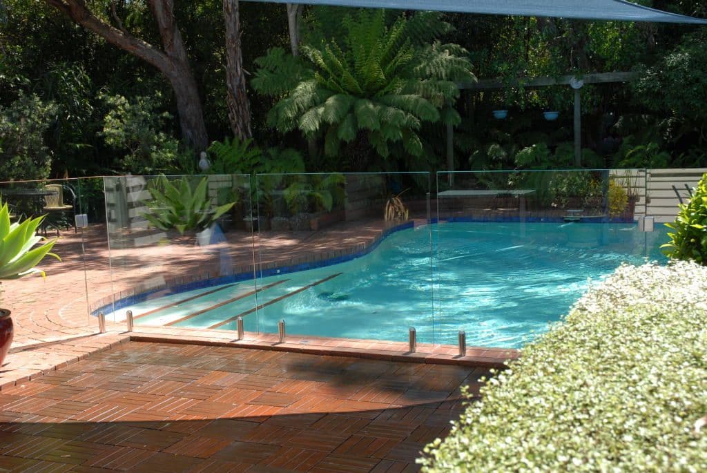 Frameless glass pool fence with chrome spigots core-drilled into brick coping surrounding a freeform backyard swimming pool in a lush garden setting.