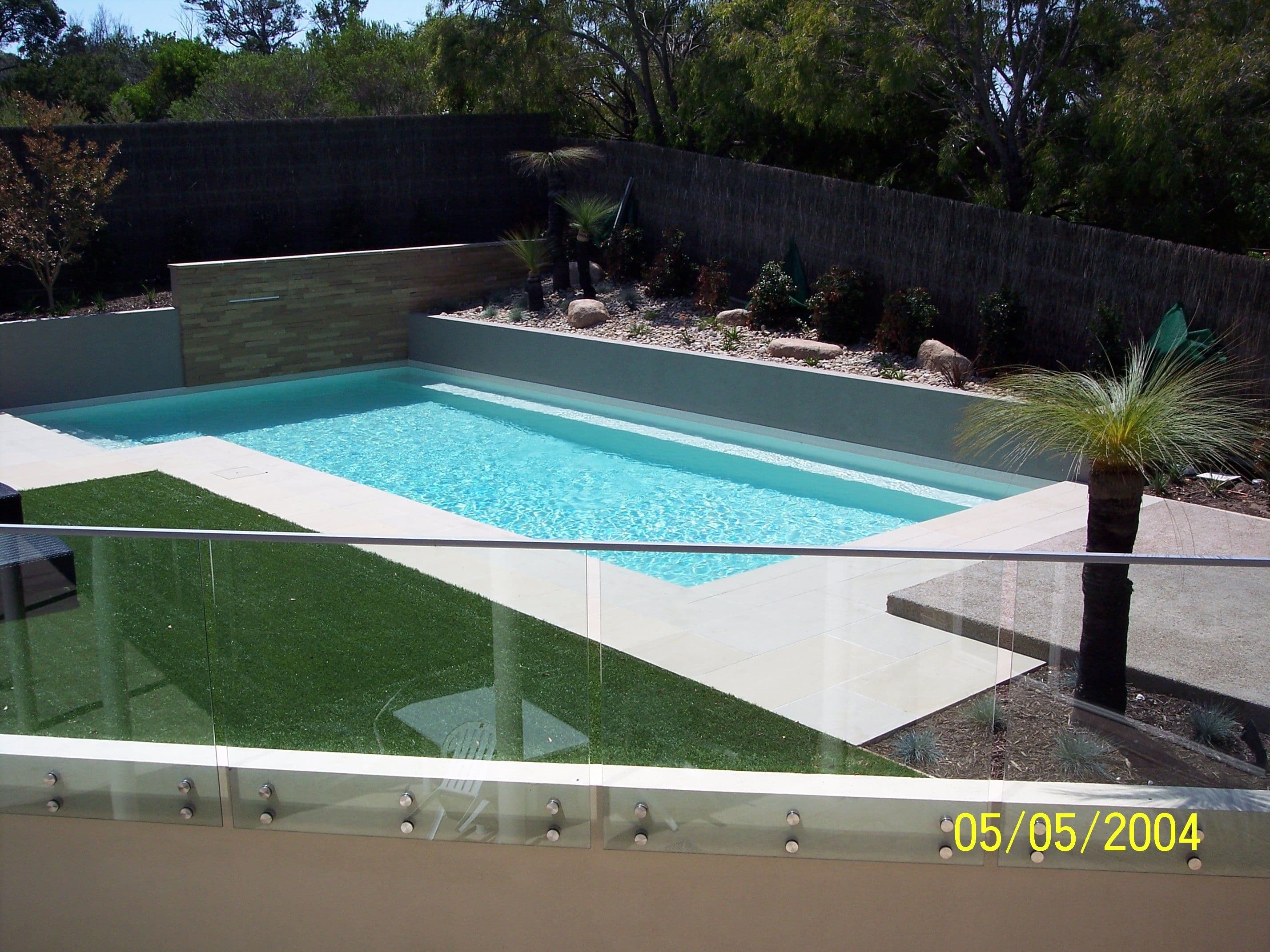 Clear glass balustrade with chrome standoff fixings mounted on a rendered wall edge overlooking a backyard pool and lawn area.