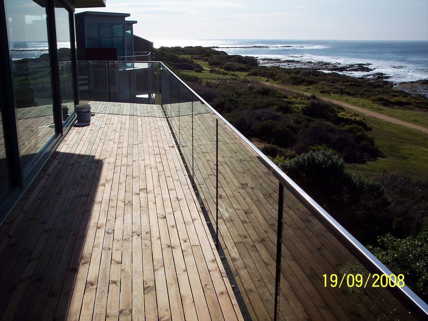 Clear glass balustrade with a chrome handrail running along a timber deck overlooking a coastal ocean view, with no pool visible.