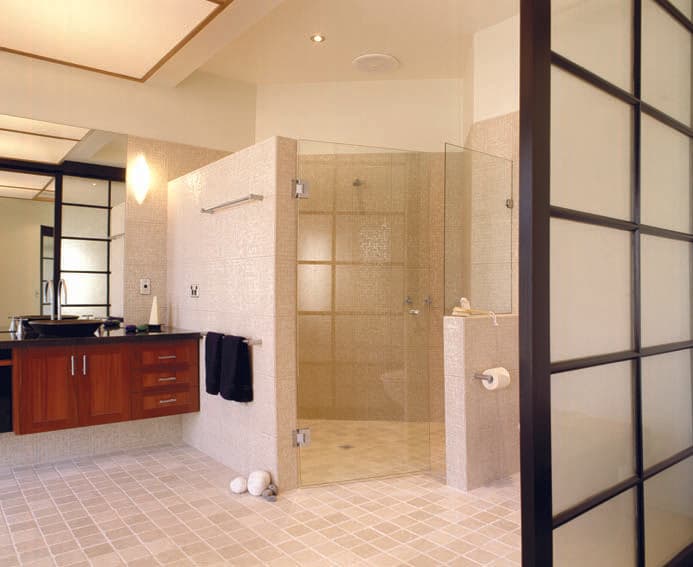 Frameless shower enclosure with a corner configuration, chrome hinges mounted directly onto the glass panels, in a neutral tiled bathroom with timber vanity cabinetry.