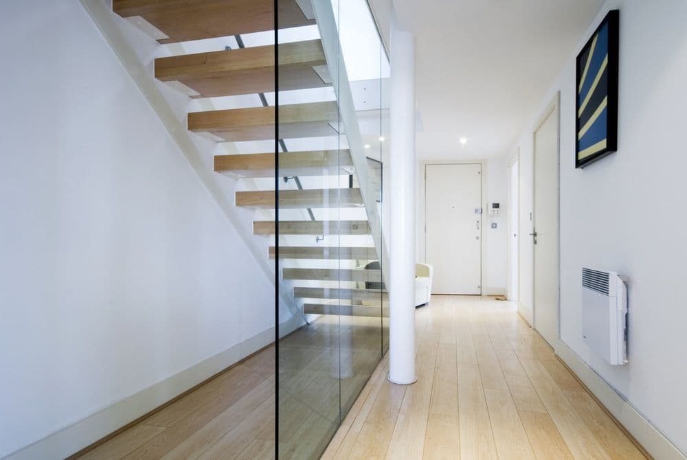 Frameless glass balustrade panel enclosing a floating timber staircase in a modern interior hallway with timber flooring.