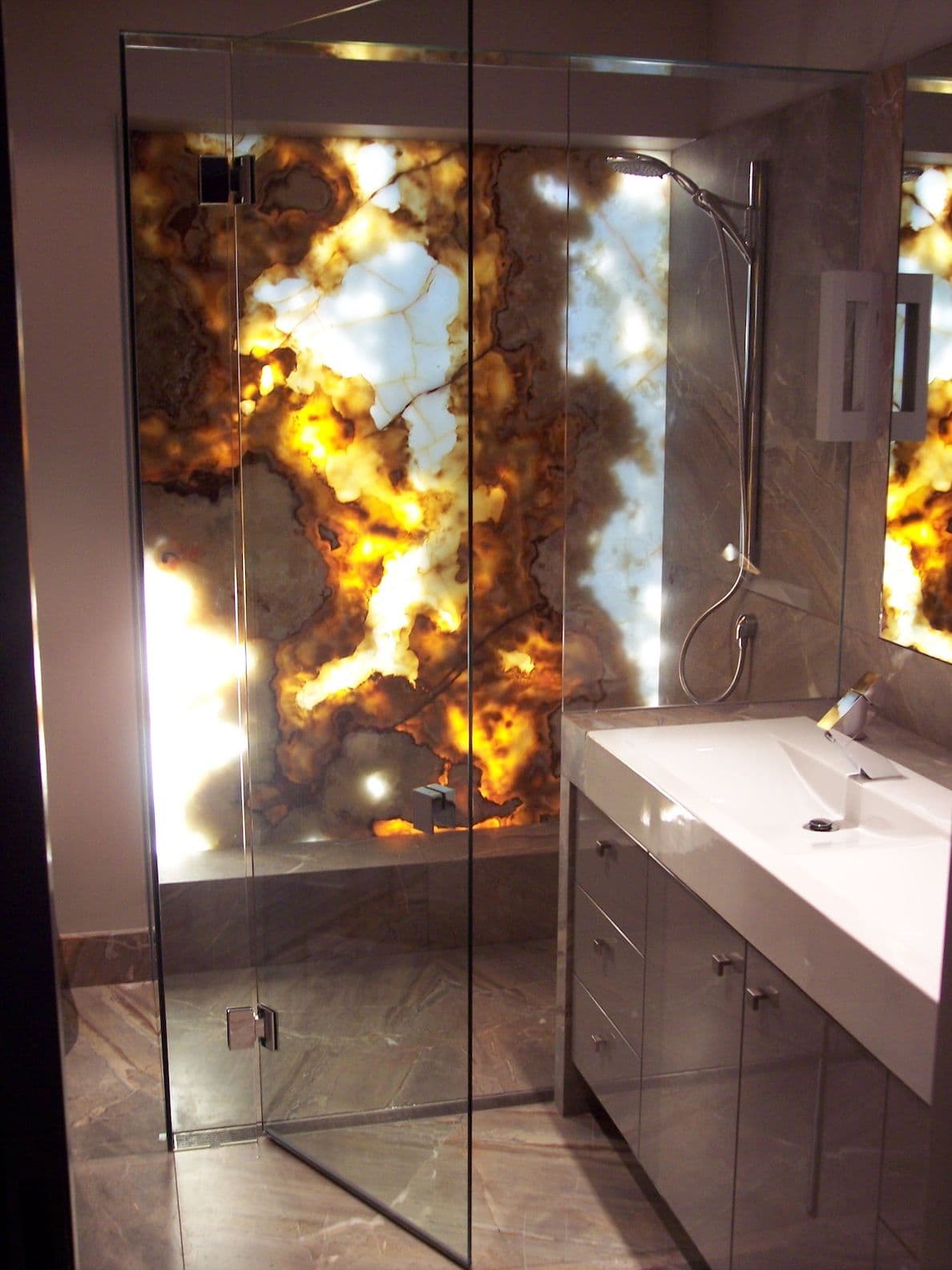 Frameless shower enclosure with clear glass panels mounted via chrome hinges directly into the glass, featuring a dramatic backlit onyx stone feature wall in a luxury marble-tiled bathroom.