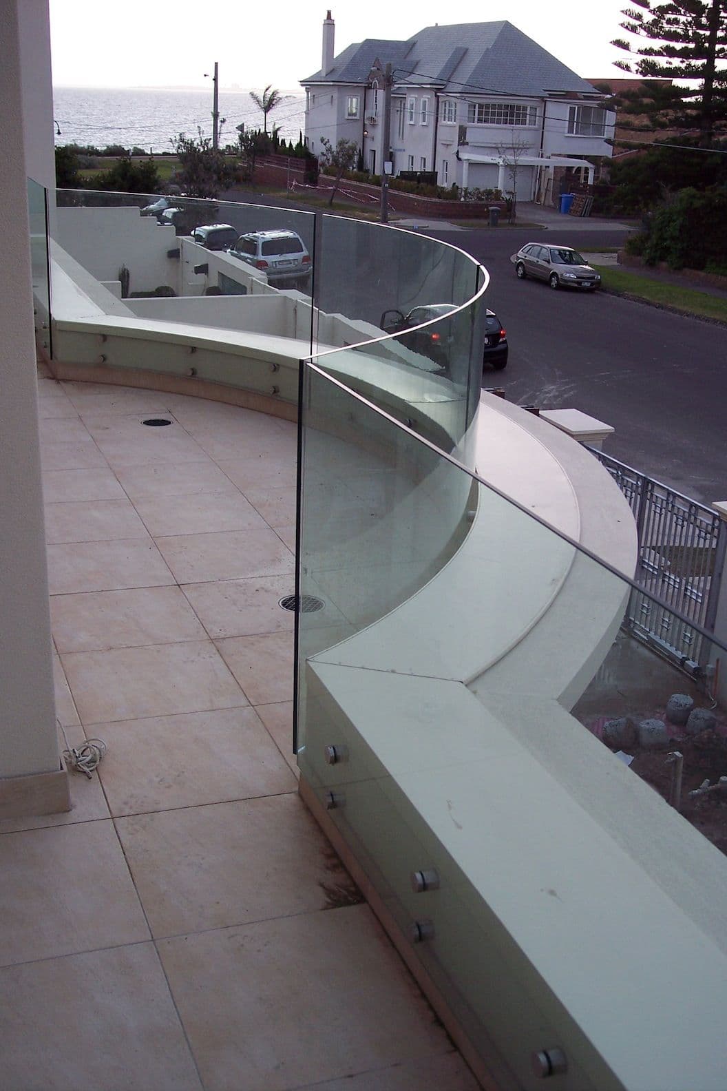 Curved frameless glass balustrade with chrome standoff spigots mounted on a rendered concrete knee wall along a tiled coastal balcony overlooking the ocean and street.
