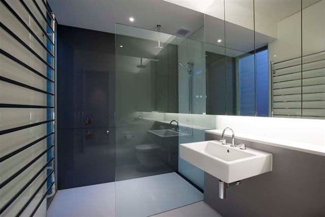 Frameless shower enclosure with clear glass panels and minimal hardware in a contemporary dark-tiled bathroom with ceiling-mounted shower head.