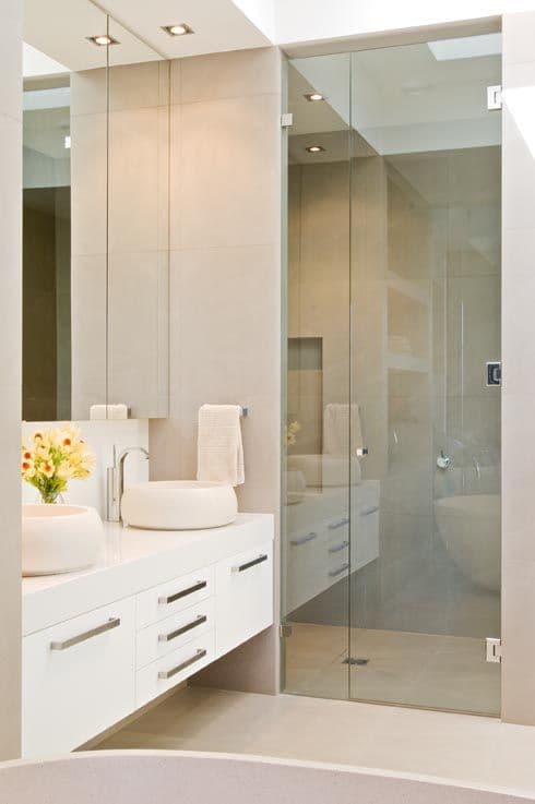Frameless shower enclosure with a hinged door and fixed panel, featuring chrome hinges and hardware mounted directly onto clear glass, in a modern white and beige tiled ensuite.