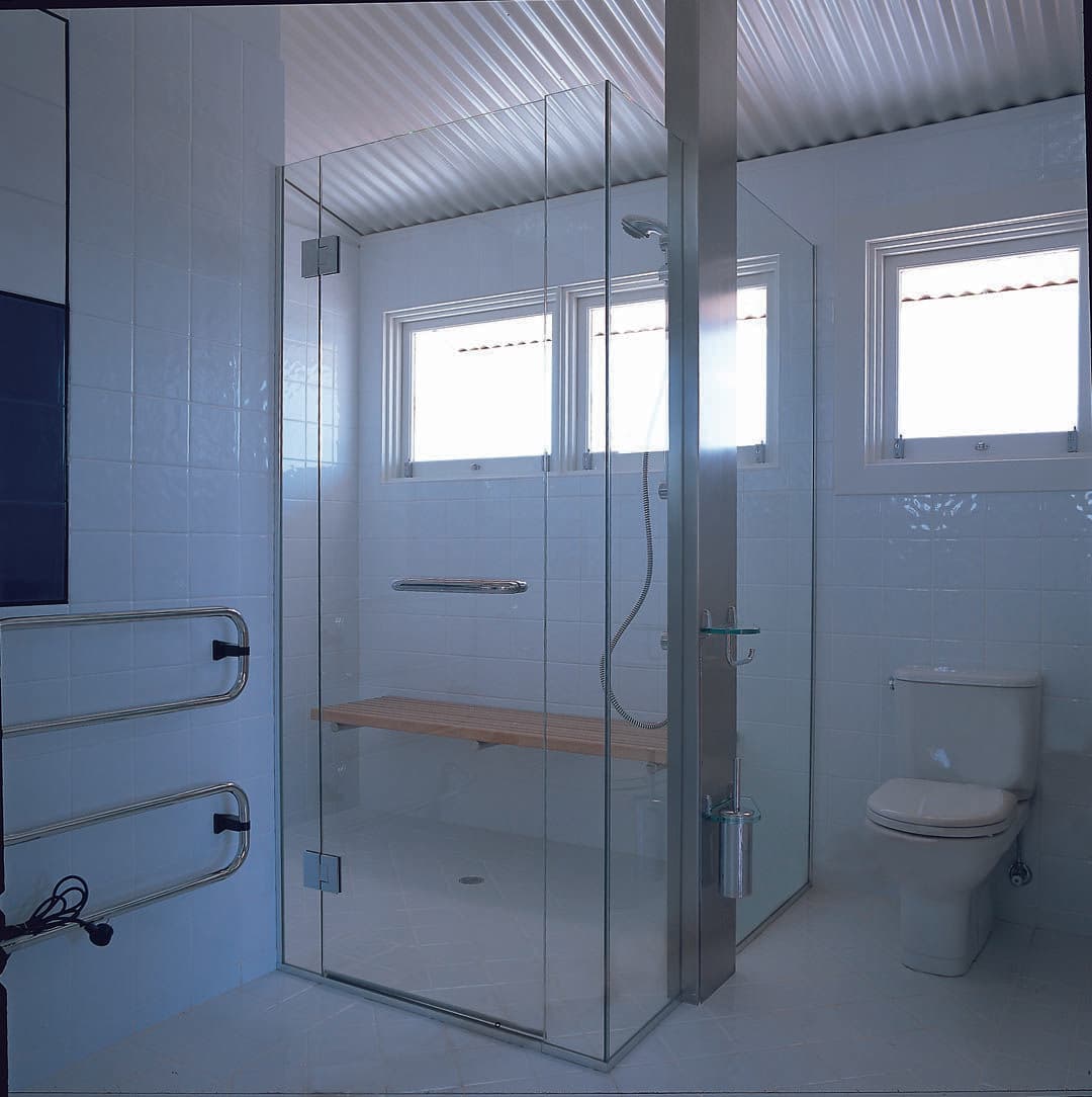 Frameless shower enclosure with chrome hinges mounted directly onto clear glass panels in a corner configuration, featuring a timber bench seat, in a white-tiled bathroom with corrugated ceiling.