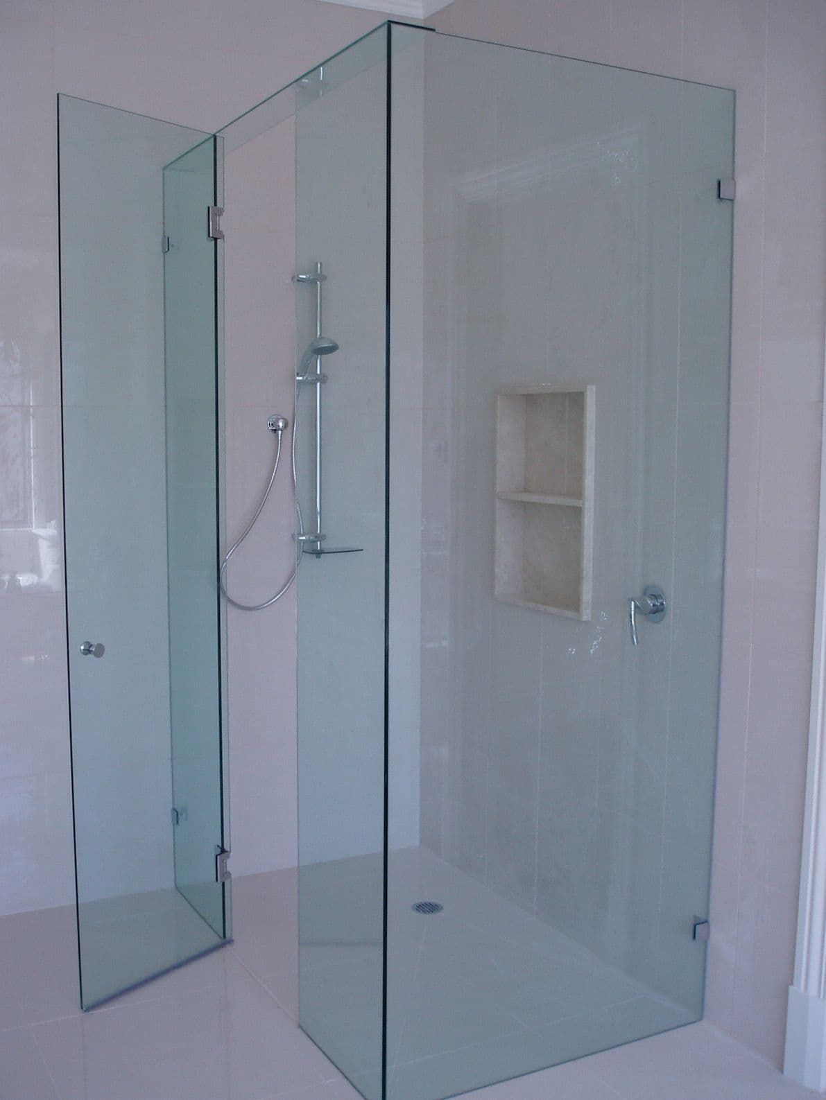 Frameless shower enclosure with a hinged door and full-height side panel, featuring chrome hinges and hardware directly mounted to clear glass, in a white tiled bathroom with a built-in niche.