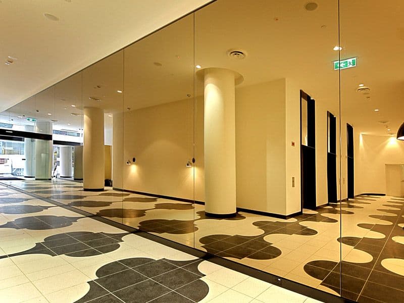 Large floor-to-ceiling mirror panels installed along a commercial corridor wall, reflecting a decorative tiled lobby interior with cylindrical columns.