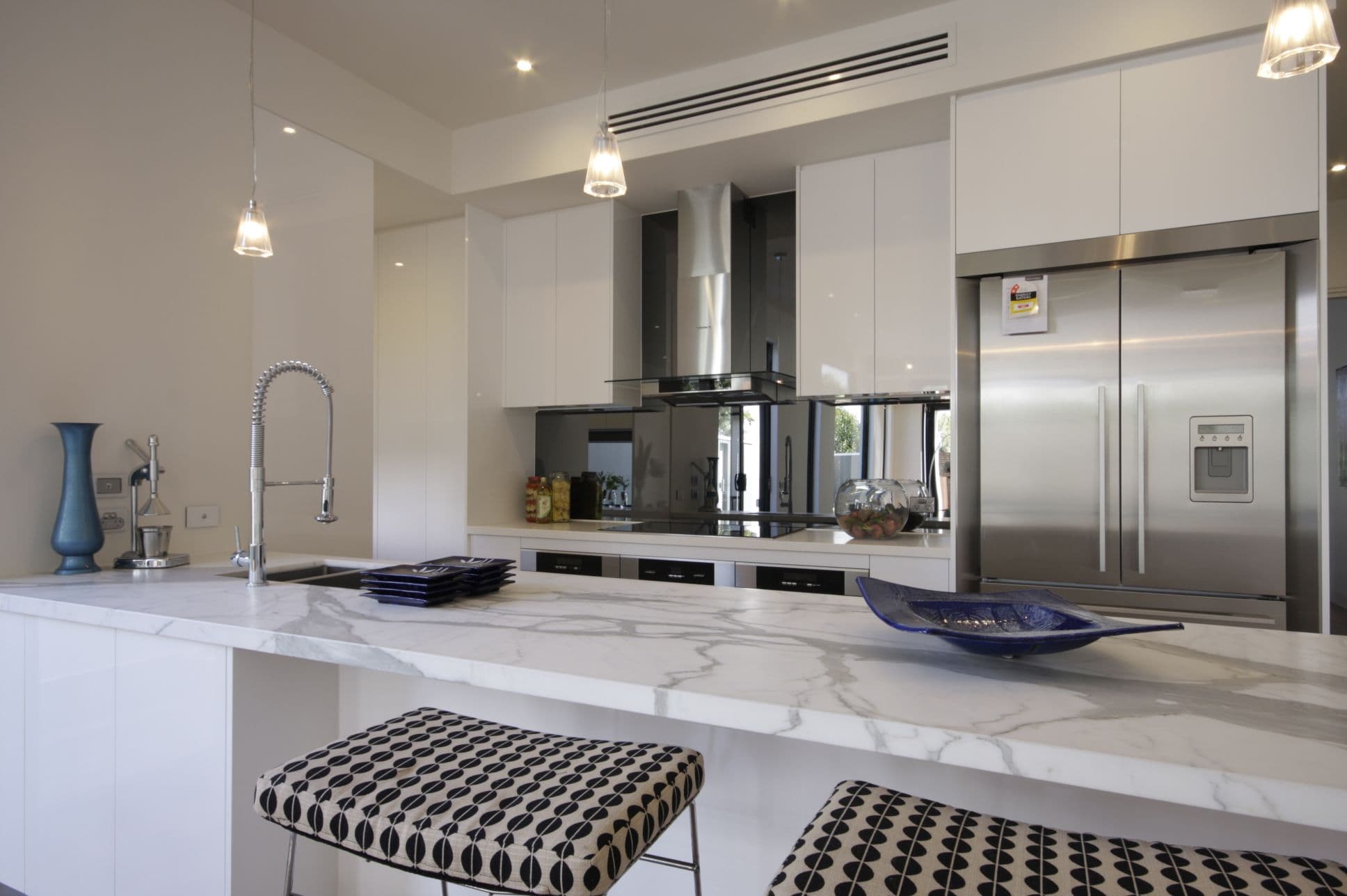 Smoke grey tinted mirror glass splashback installed behind a cooktop and range hood in a modern white kitchen with marble benchtops.