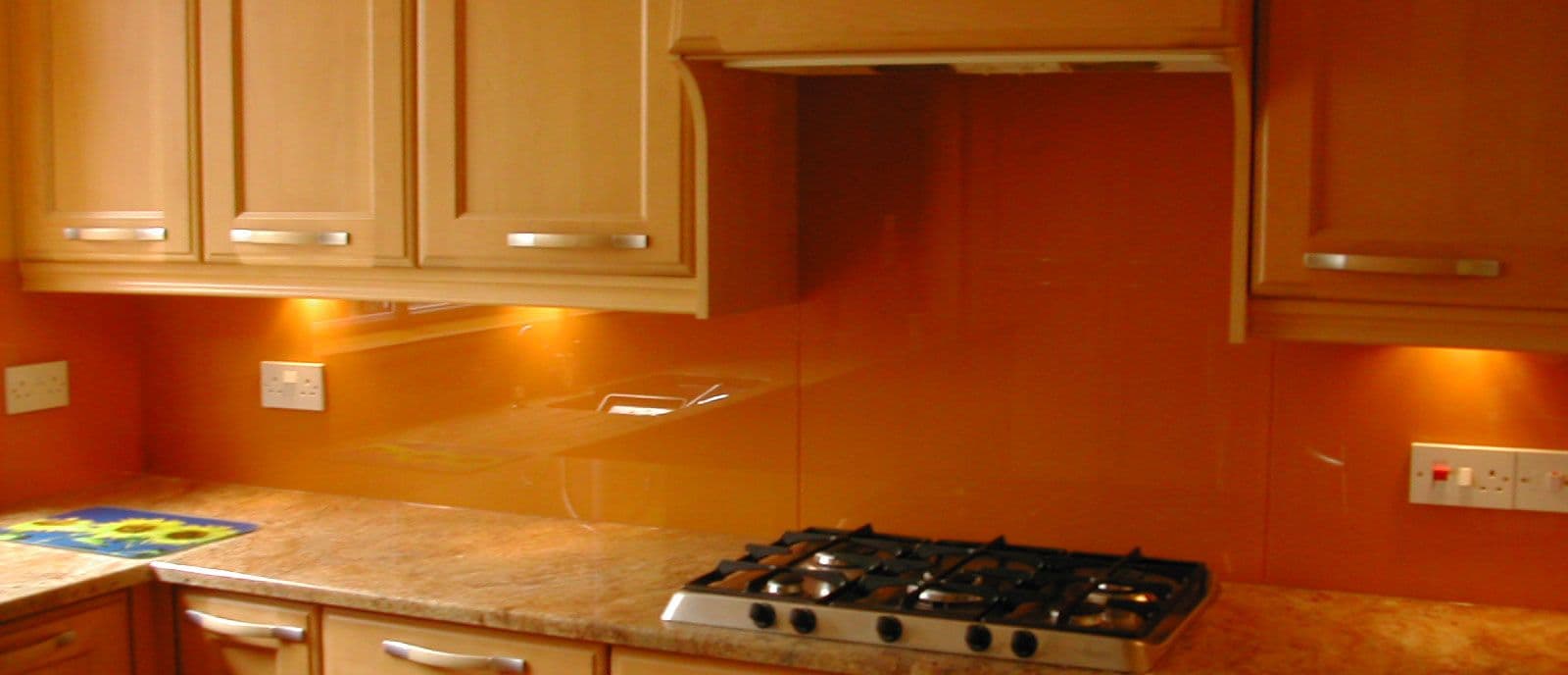Orange tinted glass splashback mounted flush on the wall behind a gas cooktop in a kitchen with timber cabinetry and stone benchtop.