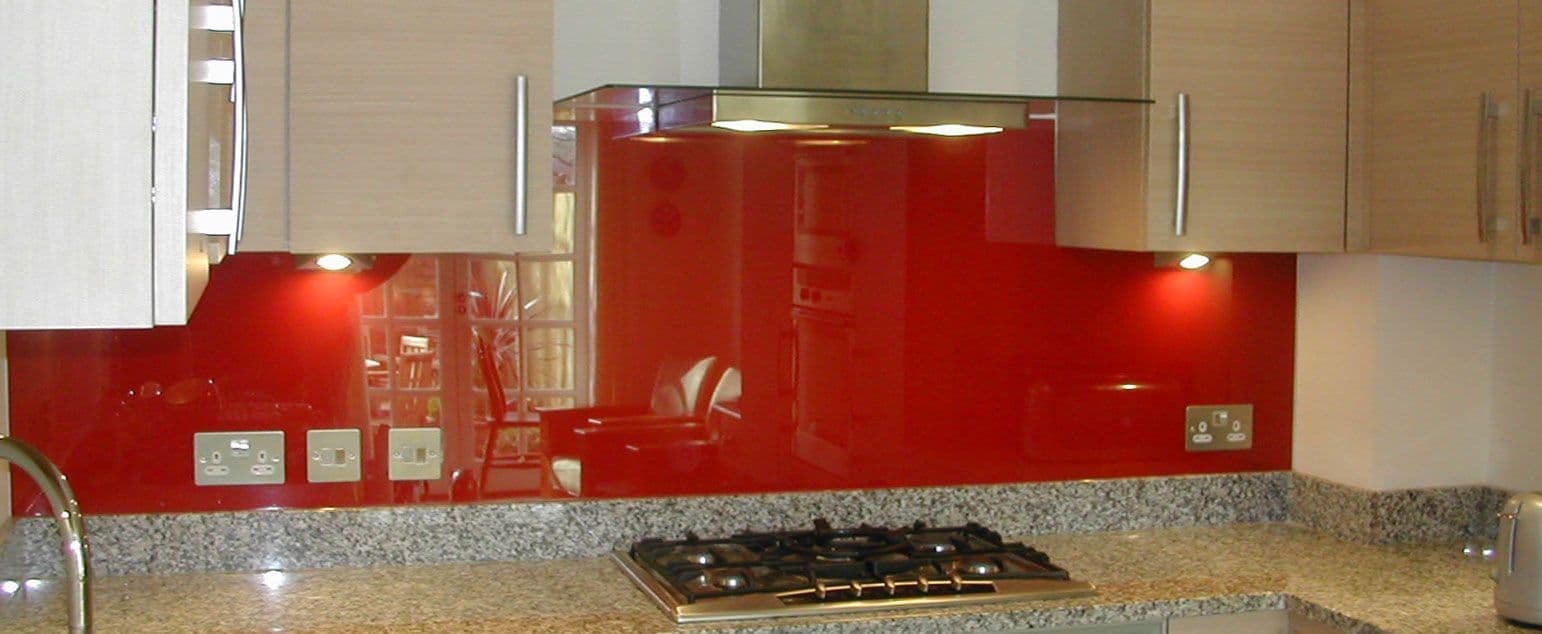 Red coloured glass splashback mounted flush behind a gas cooktop and rangehood in a modern kitchen with granite benchtops and timber cabinetry.
