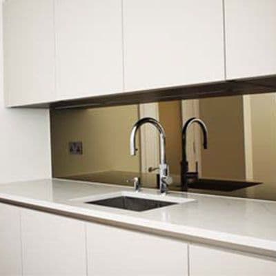 Bronze tinted mirror glass splashback installed behind a kitchen sink with chrome tapware, set between white cabinetry and a white stone benchtop.